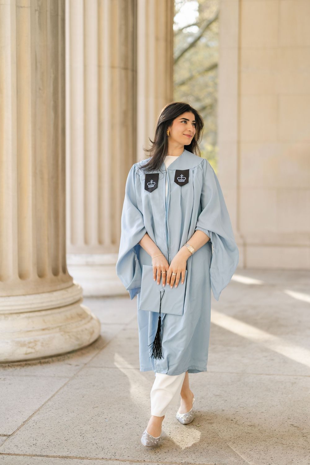 Graduate in light blue academic regalia poses between classical columns at university campus.