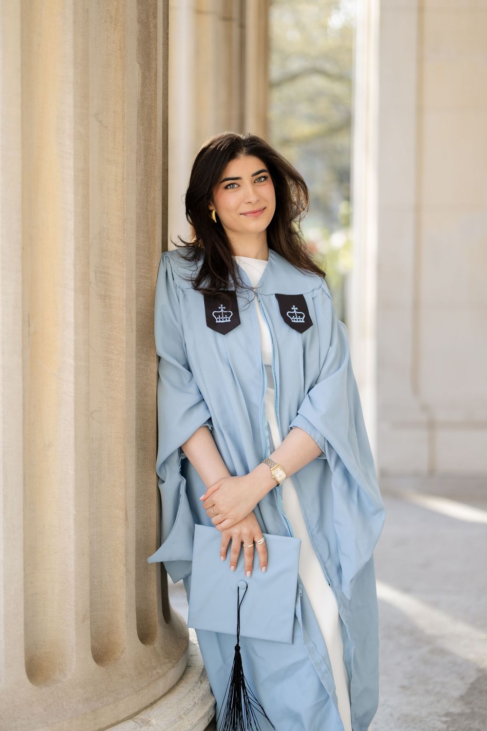 Graduate in light blue academic regalia stands in front of classic stone columns for graduation portrait.