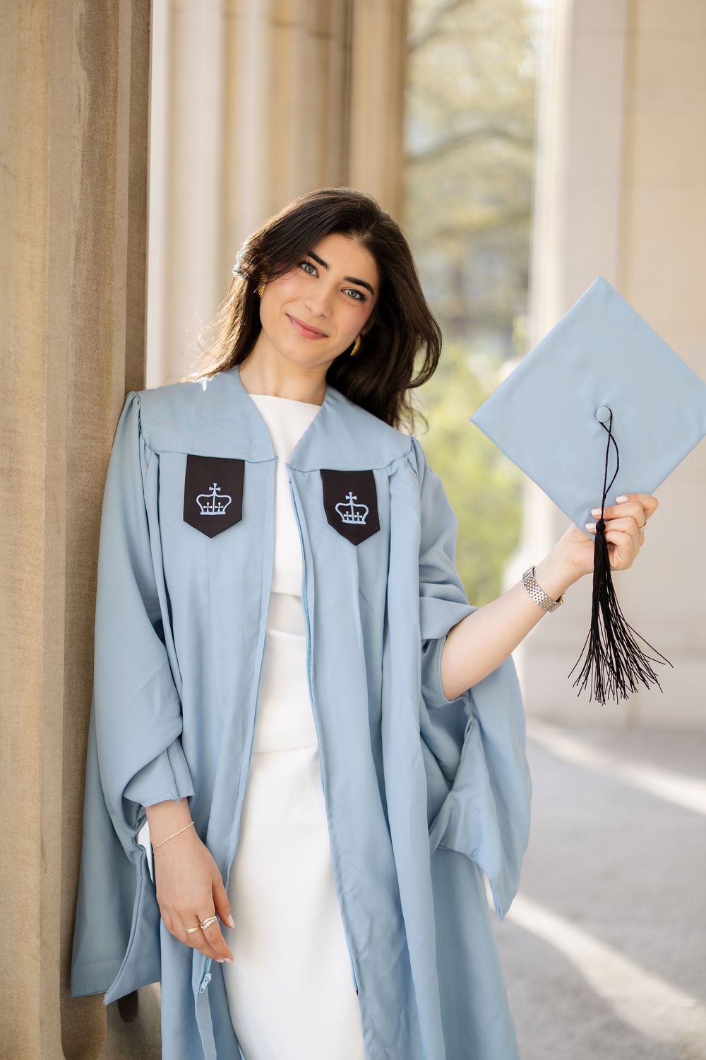 Graduate in light blue academic regalia holds up diploma and tassel against columned building backdrop.