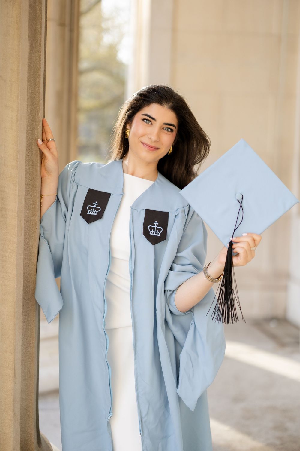 Graduate student in light blue academic regalia holding diploma and tassel while posing against marble column.