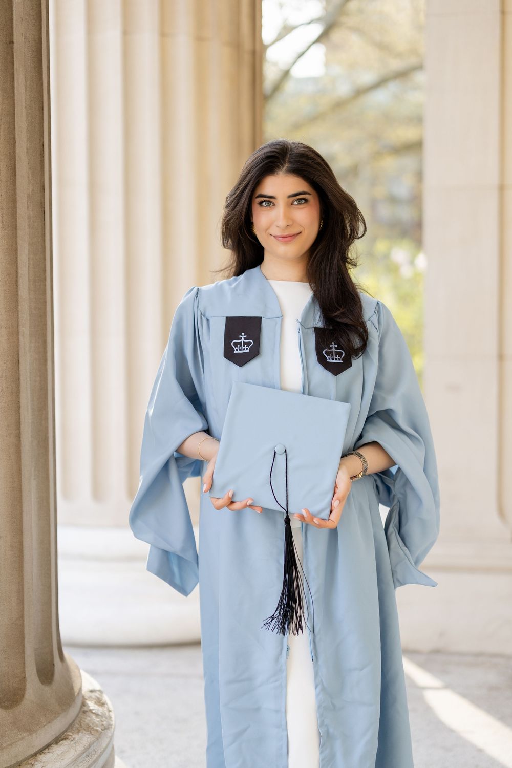 A graduate in light blue academic regalia holds their diploma while standing between classical stone columns.