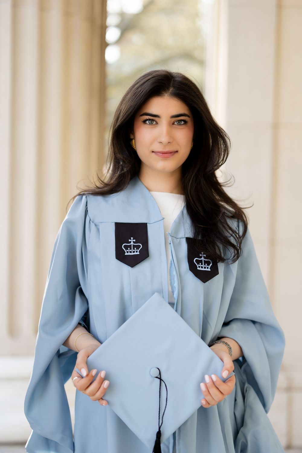 Graduate in light blue academic regalia holds matching graduation cap near white stone columns.