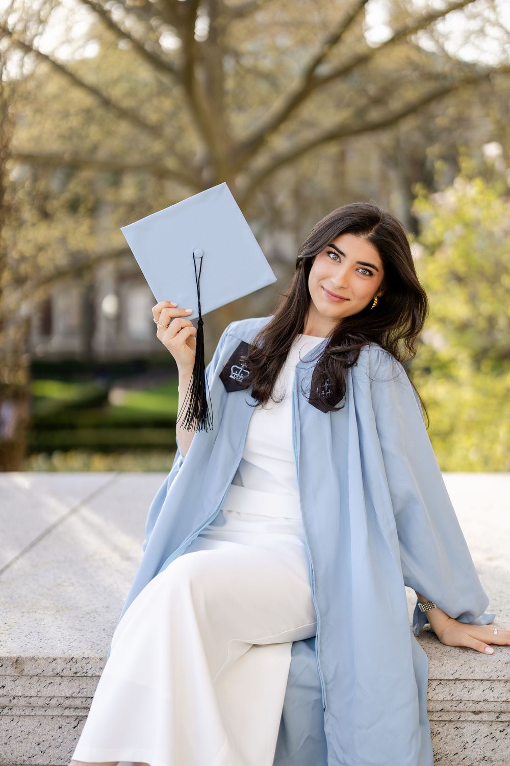 Graduate in light blue coat sits on concrete steps holding diploma on sunny day outdoors.