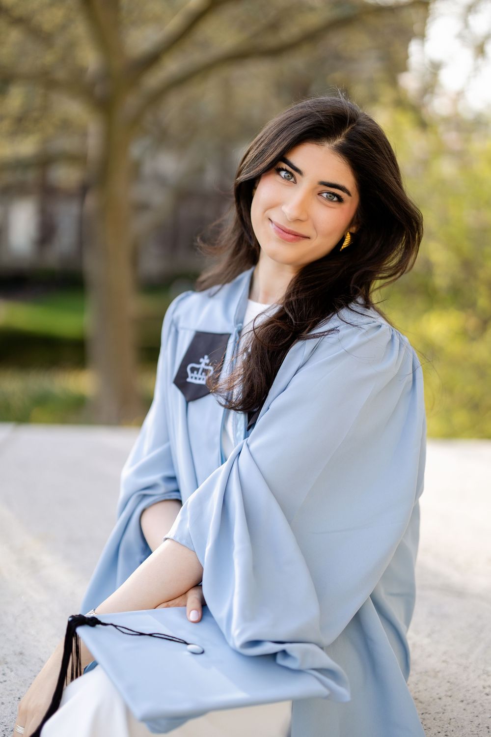 Graduate in light blue graduation gown sits outdoors on sunny spring day with trees in background.