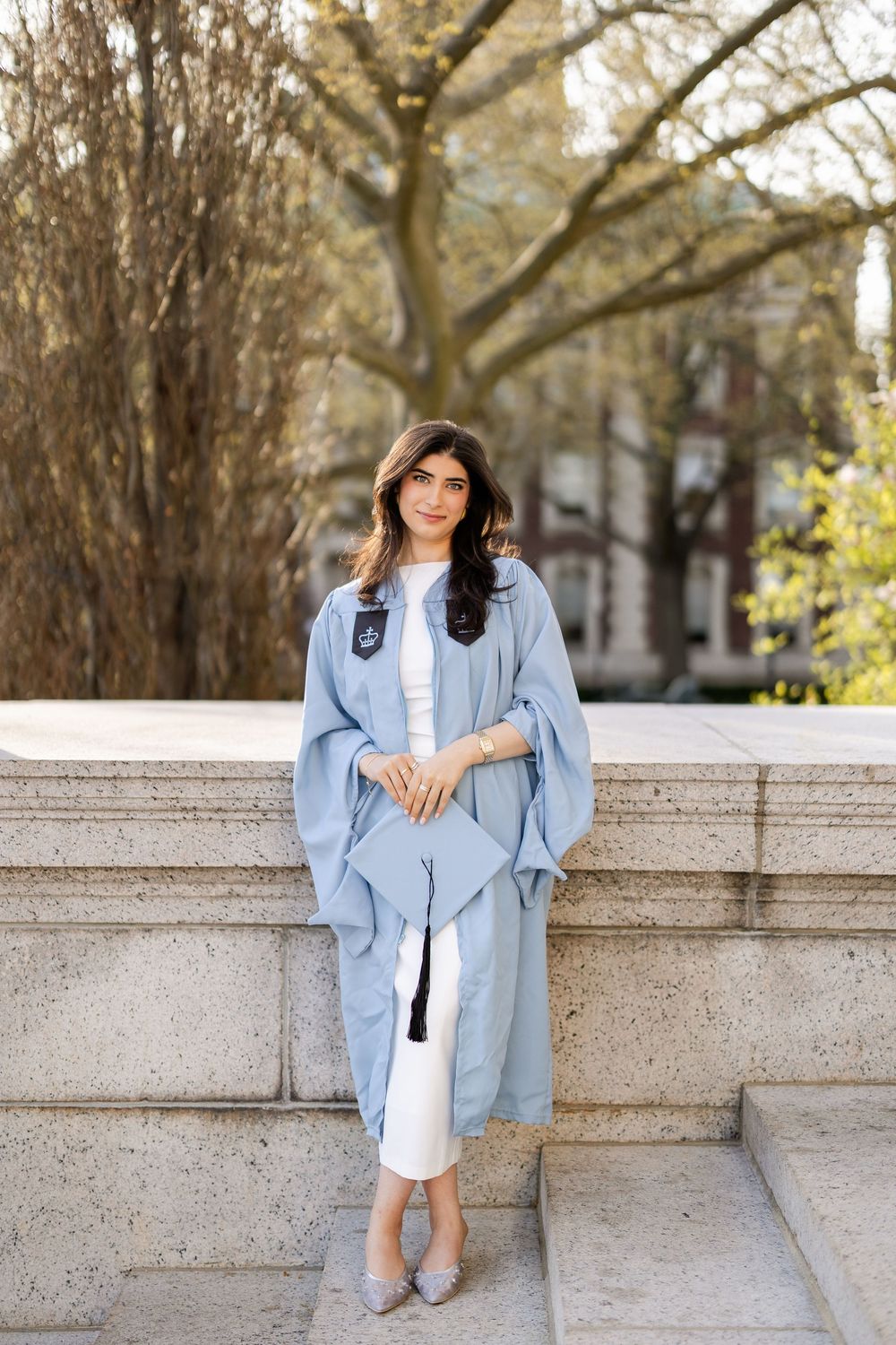Graduate wearing light blue academic regalia poses on stone steps during graduation ceremony on campus.
