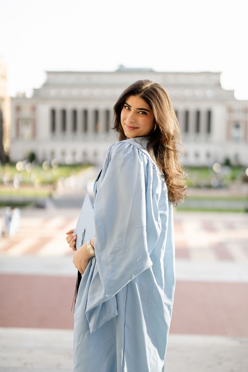 Graduate in light blue commencement gown stands in front of historic columned building on sunny day.