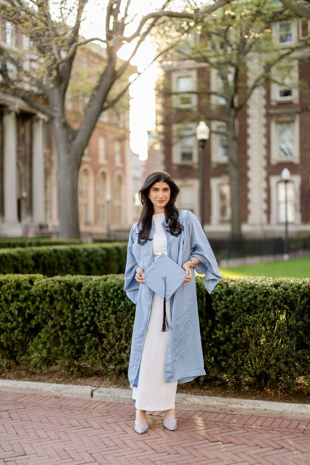 Person in a light blue long coat and white pants poses on brick path near classic brick buildings and manicured hedges.