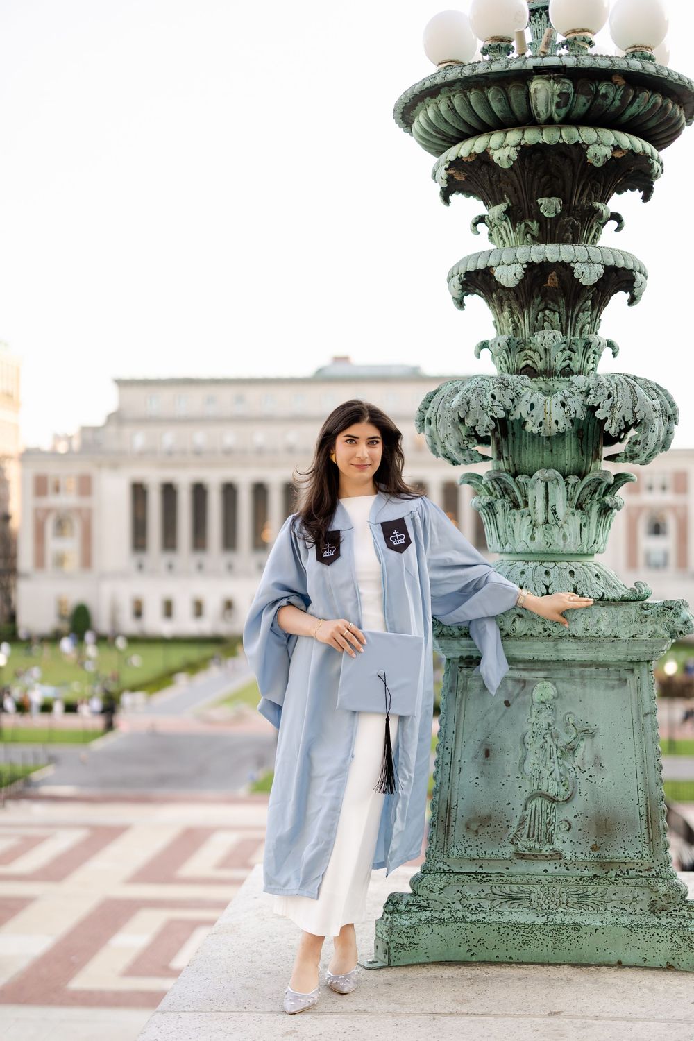 Graduate in light blue academic gown poses next to ornate vintage lamp post on campus grounds.
