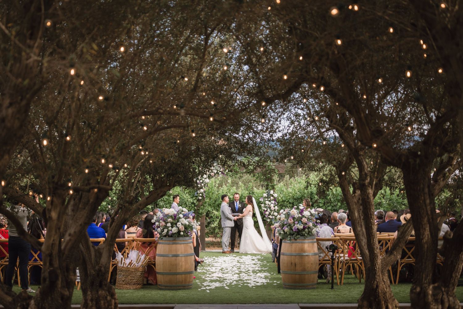 Rustic outdoor wedding ceremony under trees with wine barrels and string lights creating a romantic atmosphere.