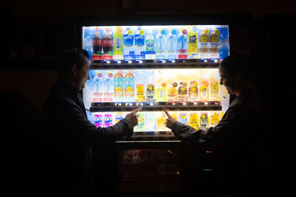 Two people browse a brightly lit Japanese vending machine together in the dark.