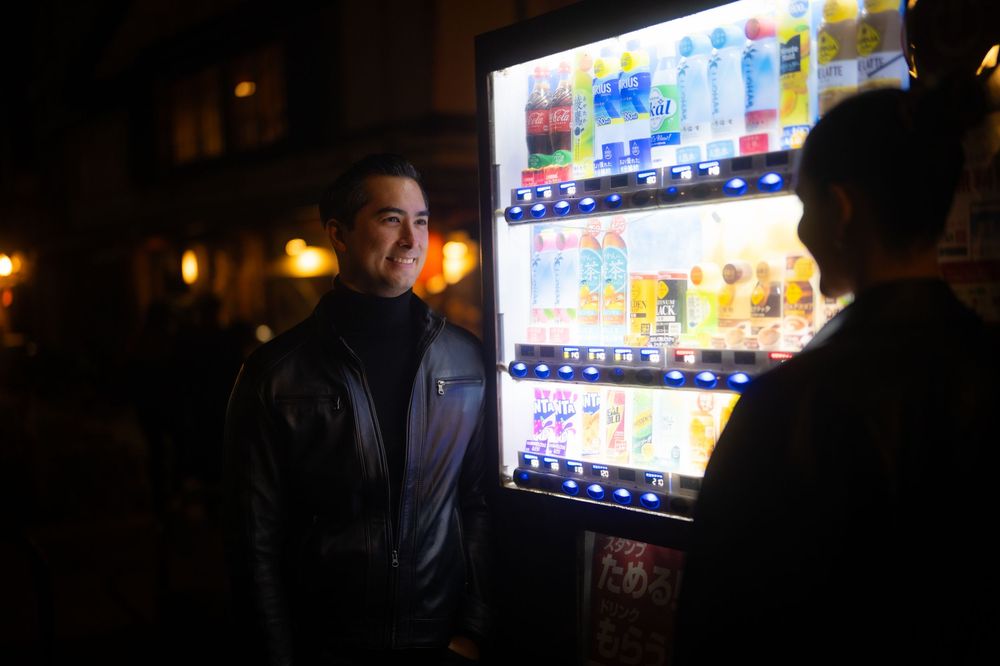 A smiling man in a black jacket stands beside a glowing Japanese vending machine at night.