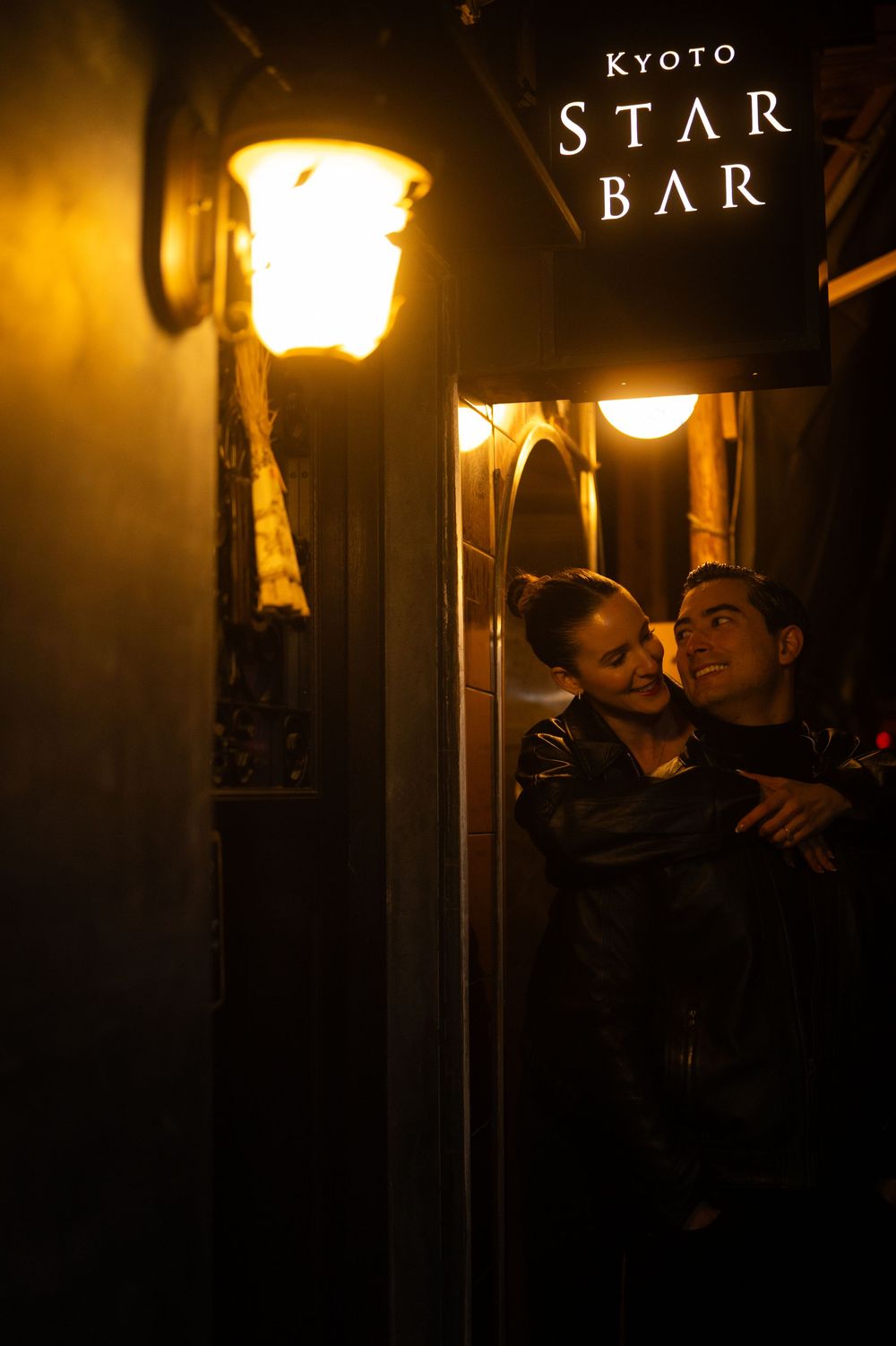 A couple embraces warmly outside the Kyoto Star Bar beneath a glowing golden lantern at night.