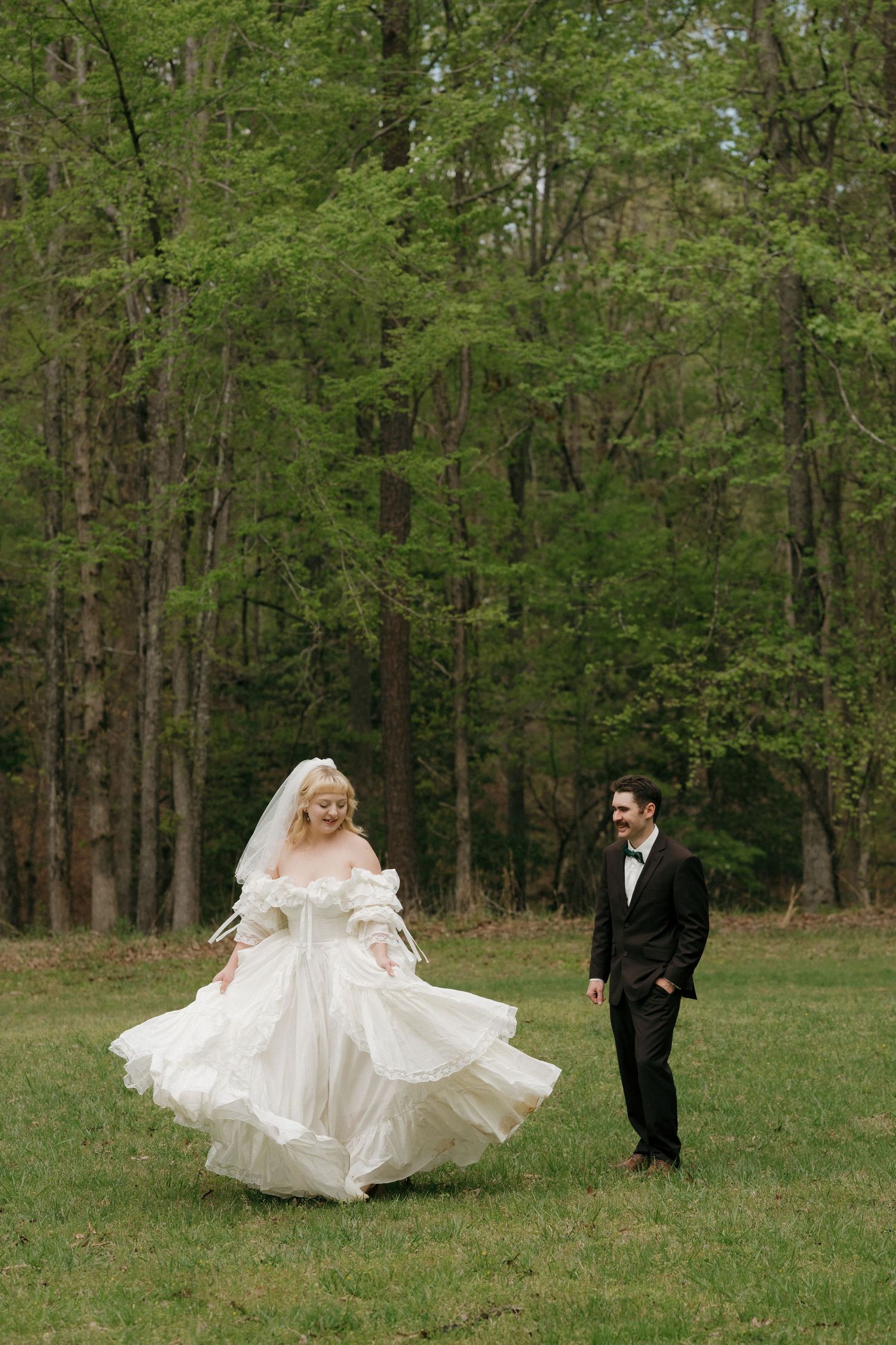 A couple in wedding attire walks through a lush green forest setting.
