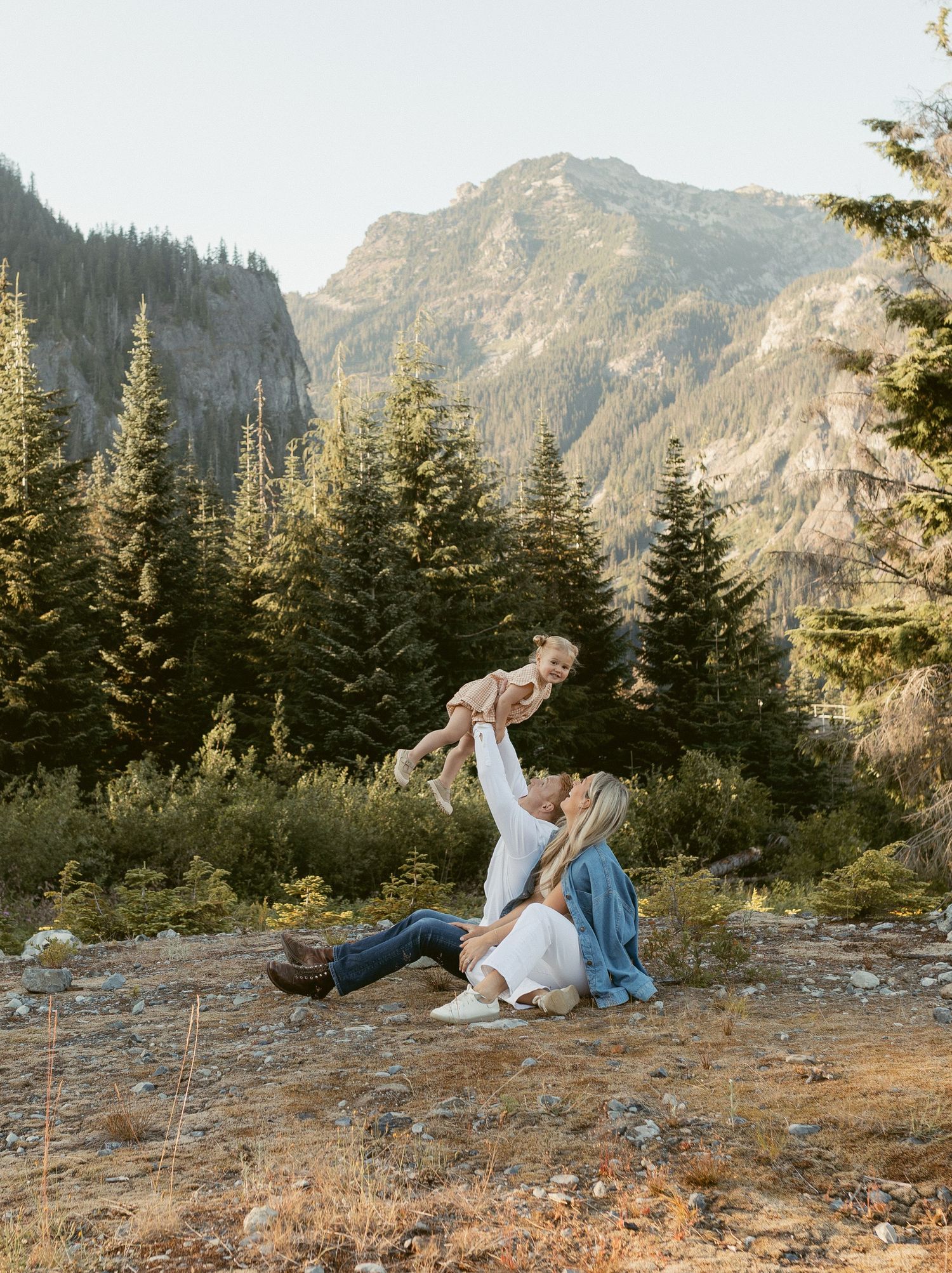 A couple sits together on a rocky mountain overlook surrounded by evergreen trees and rugged peaks at sunset.