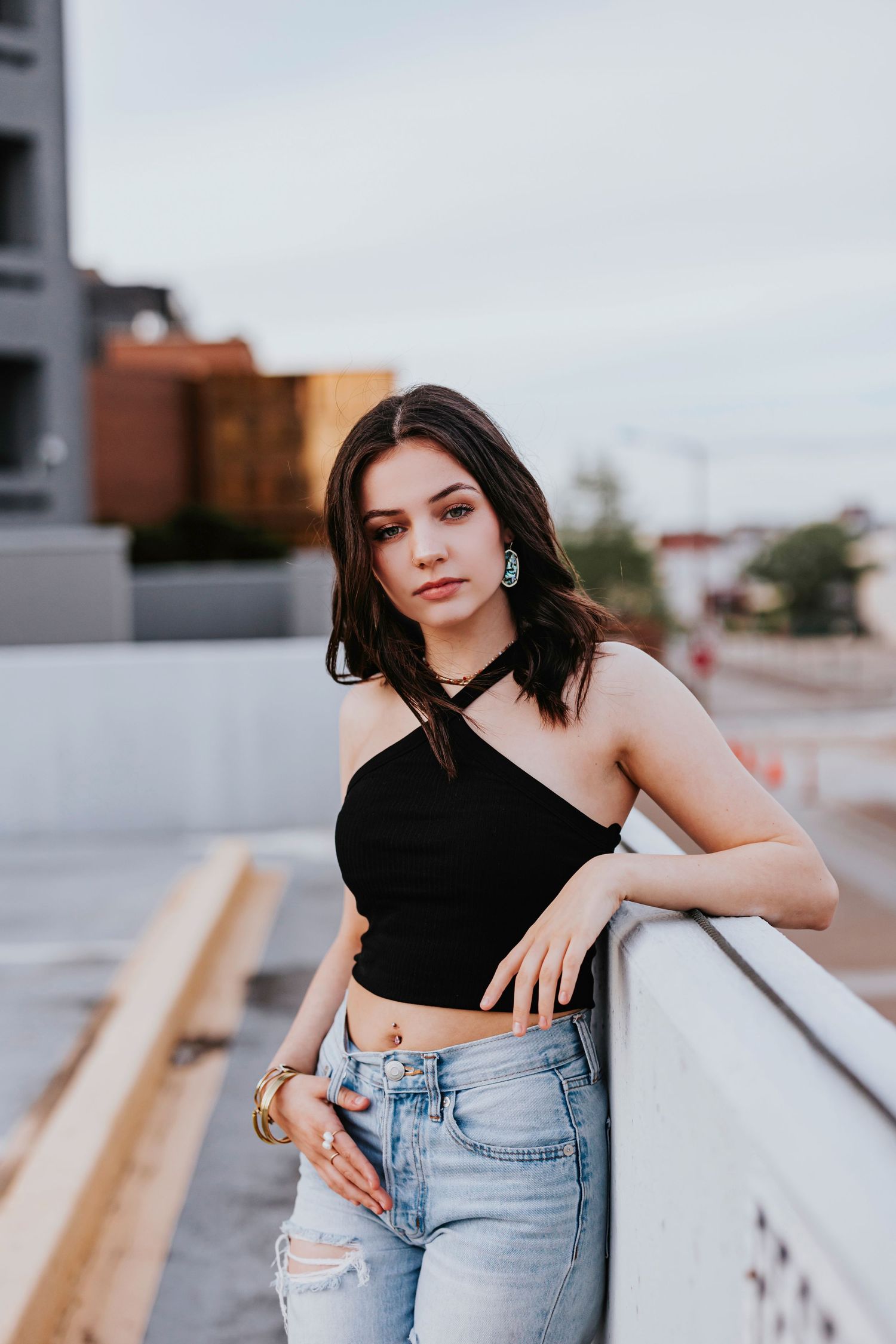 Woman in black crop top and jeans poses on rooftop during urban photoshoot.
