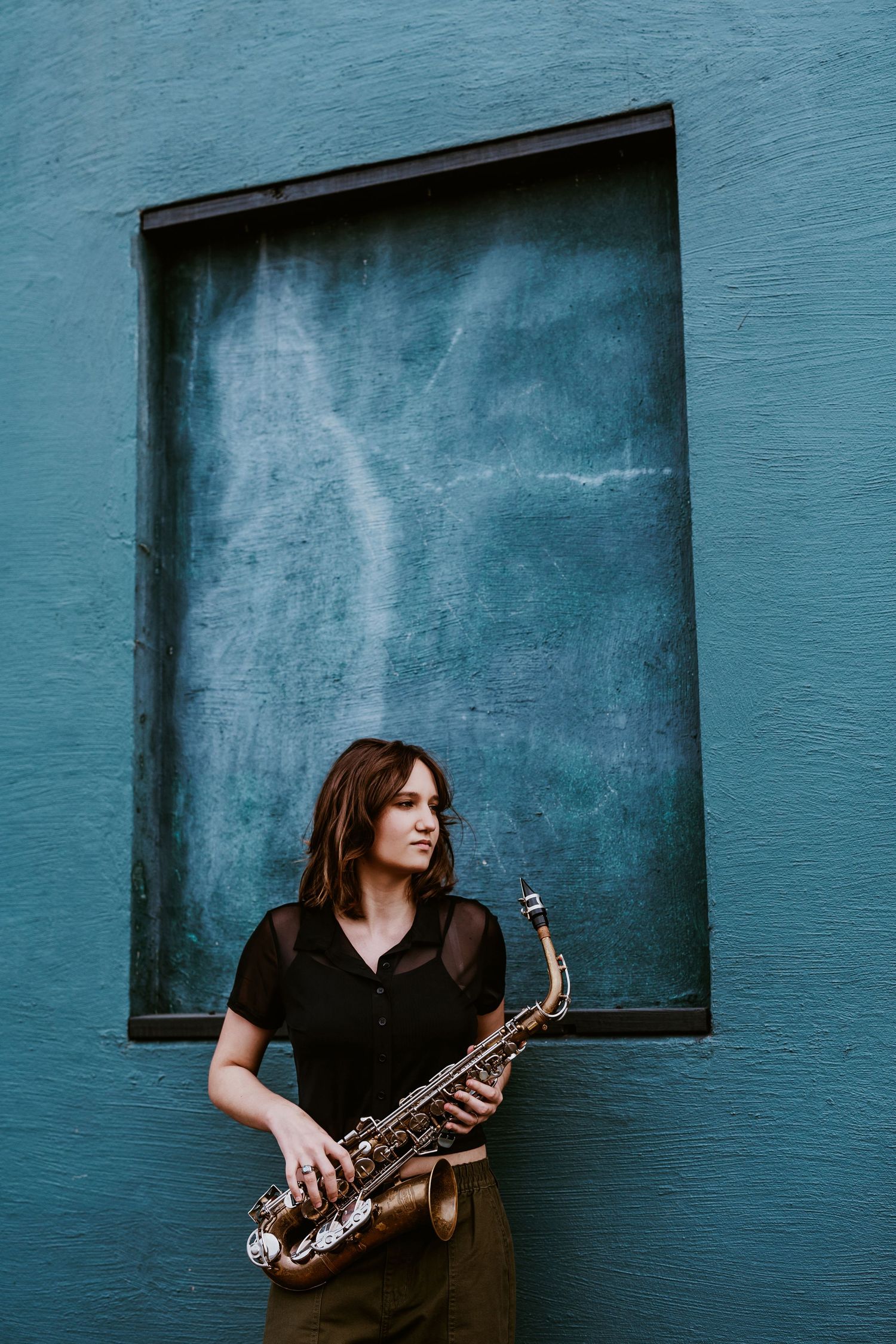 Musician poses with saxophone against turquoise wall with dramatic lighting.