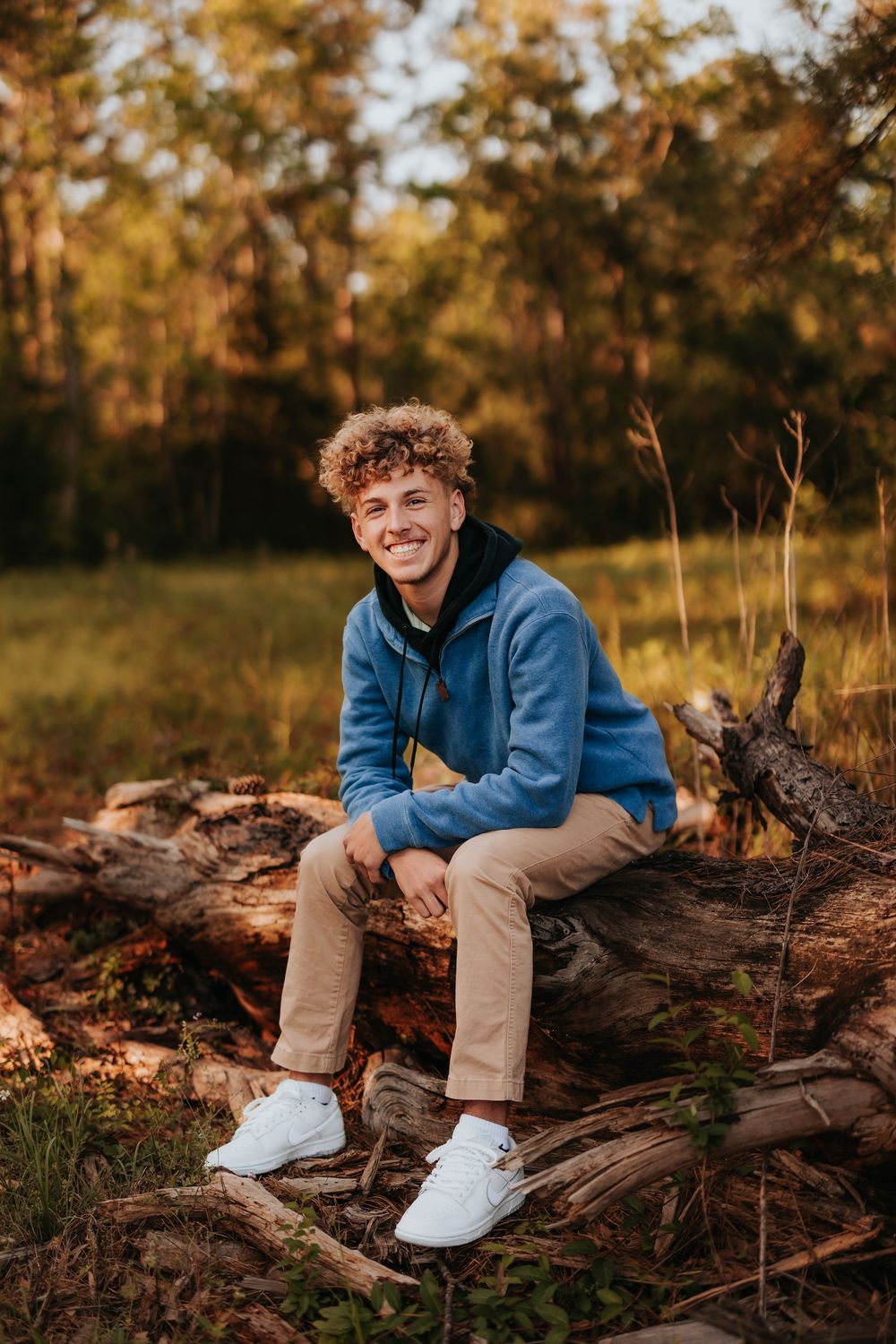Person sits casually on fallen logs wearing blue hoodie and khakis during autumn sunset.