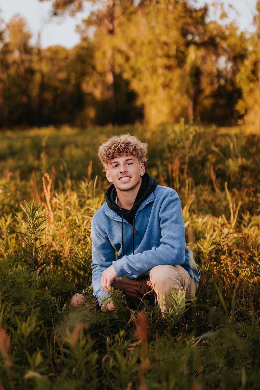 Person in blue sweatshirt sits in golden grass during sunset photoshoot.