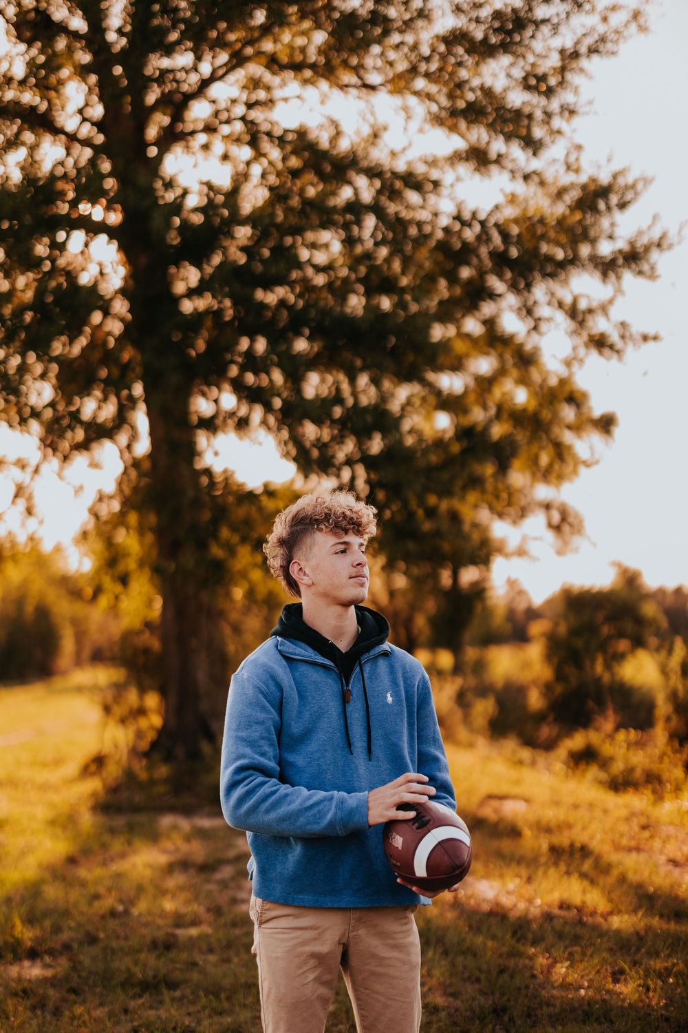 Person holds football while sitting outdoors during golden hour wearing blue hoodie.