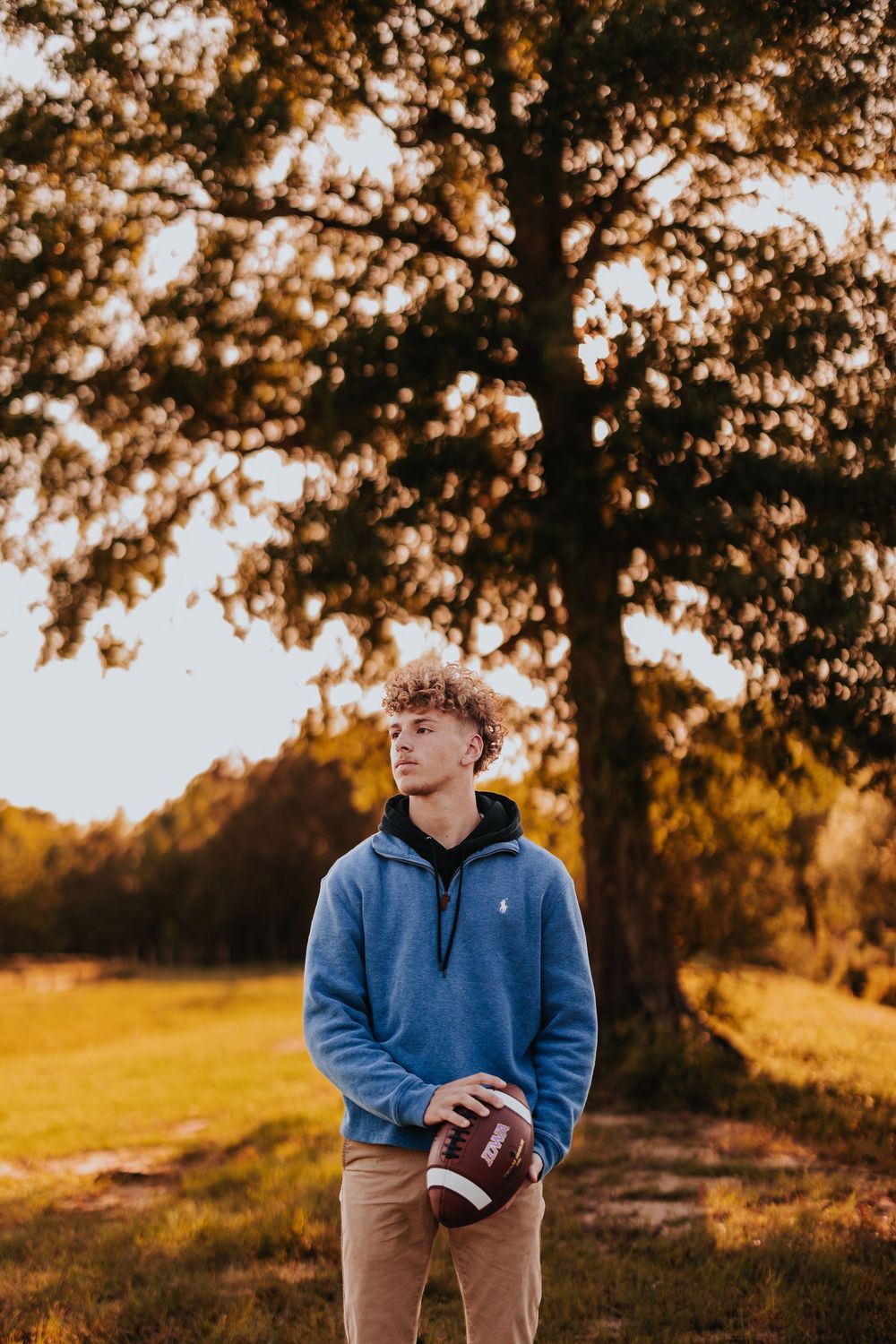 Person poses with football against autumn trees during sunset wearing blue casual attire.