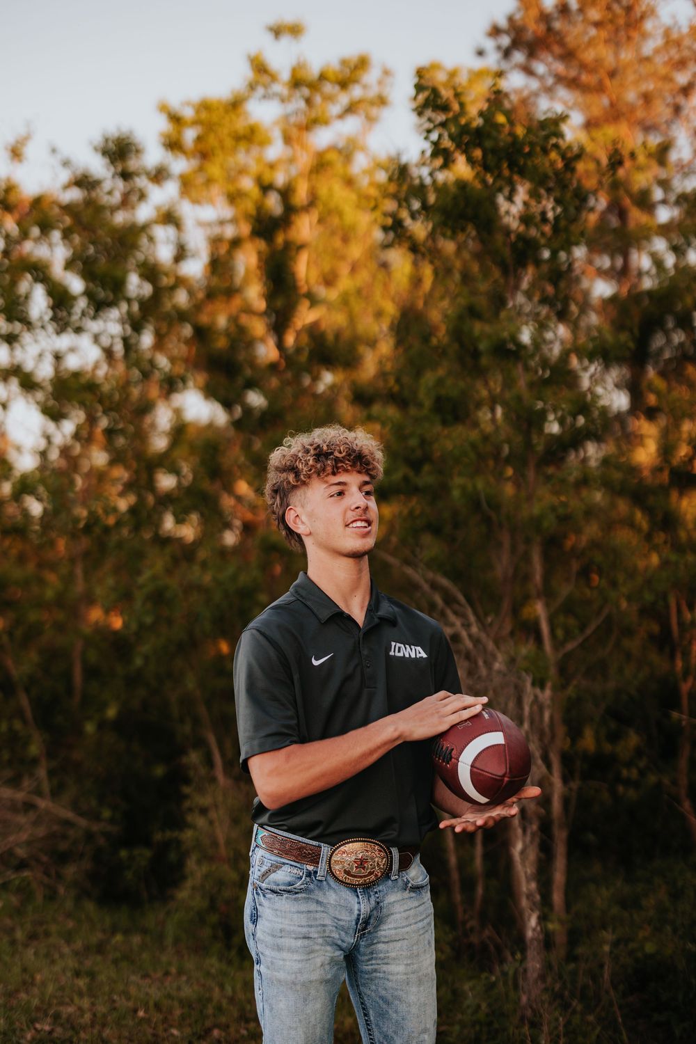 Person in black Nike shirt and jeans poses playfully with football at sunset.