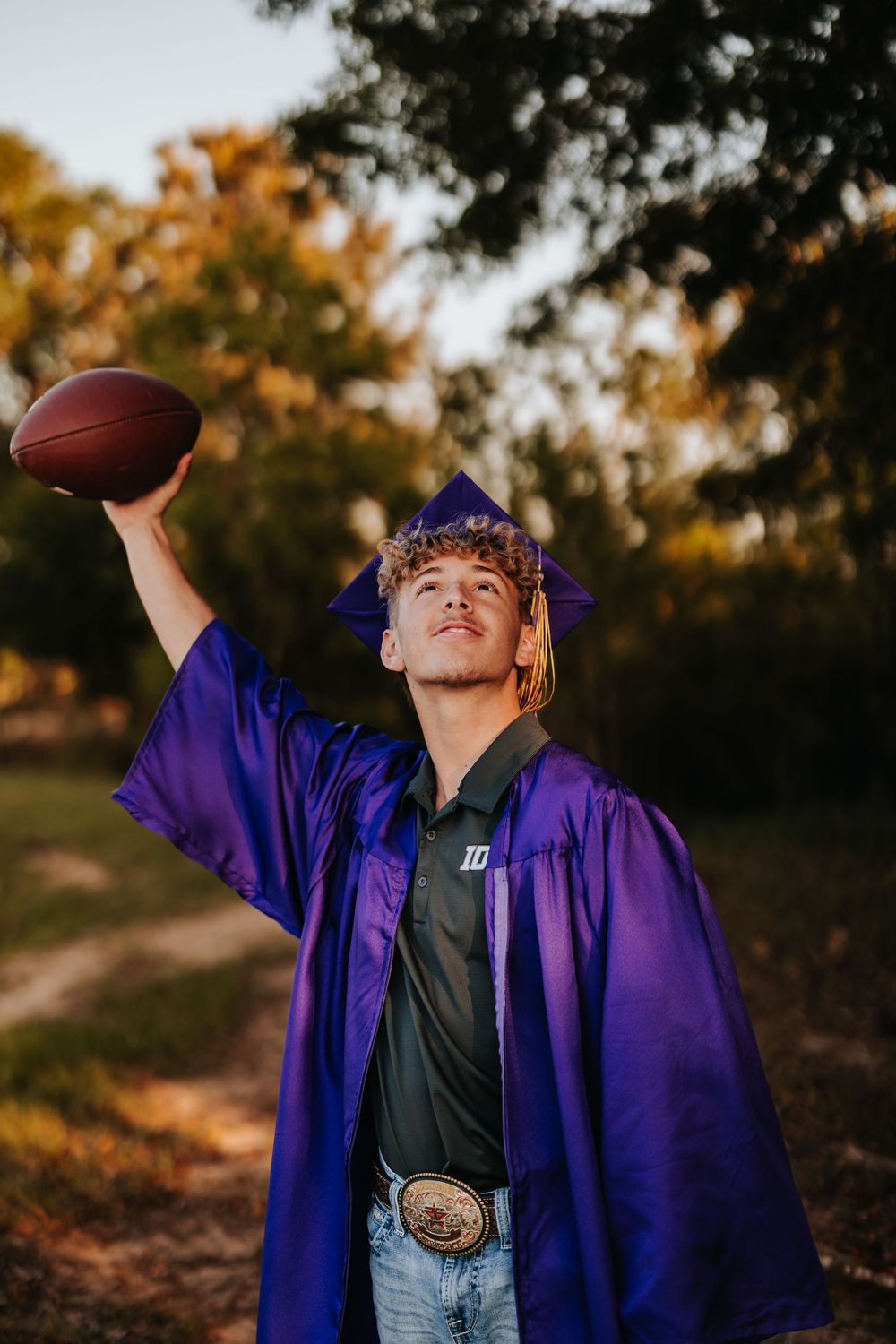 Person wearing purple graduation robe throws football in outdoor autumn setting.