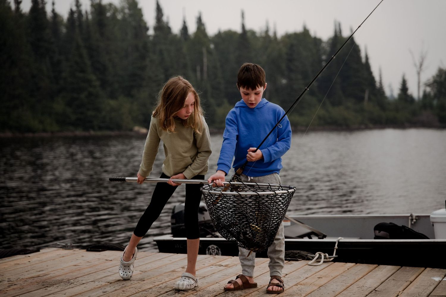 People fishing from a wooden dock on a tranquil lake surrounded by evergreen trees.
