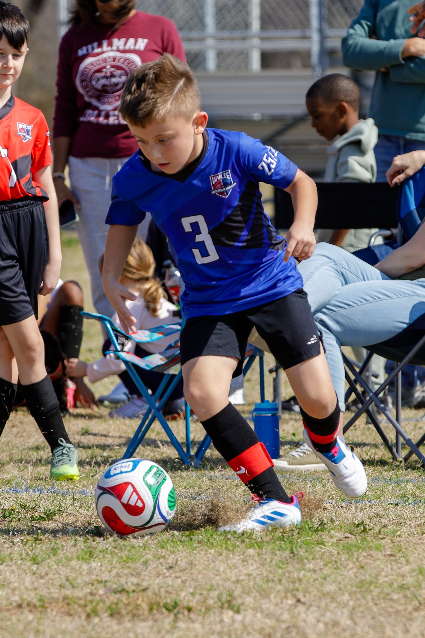 Youth soccer players in blue and red uniforms sit on small chairs during a game break on a grassy field.