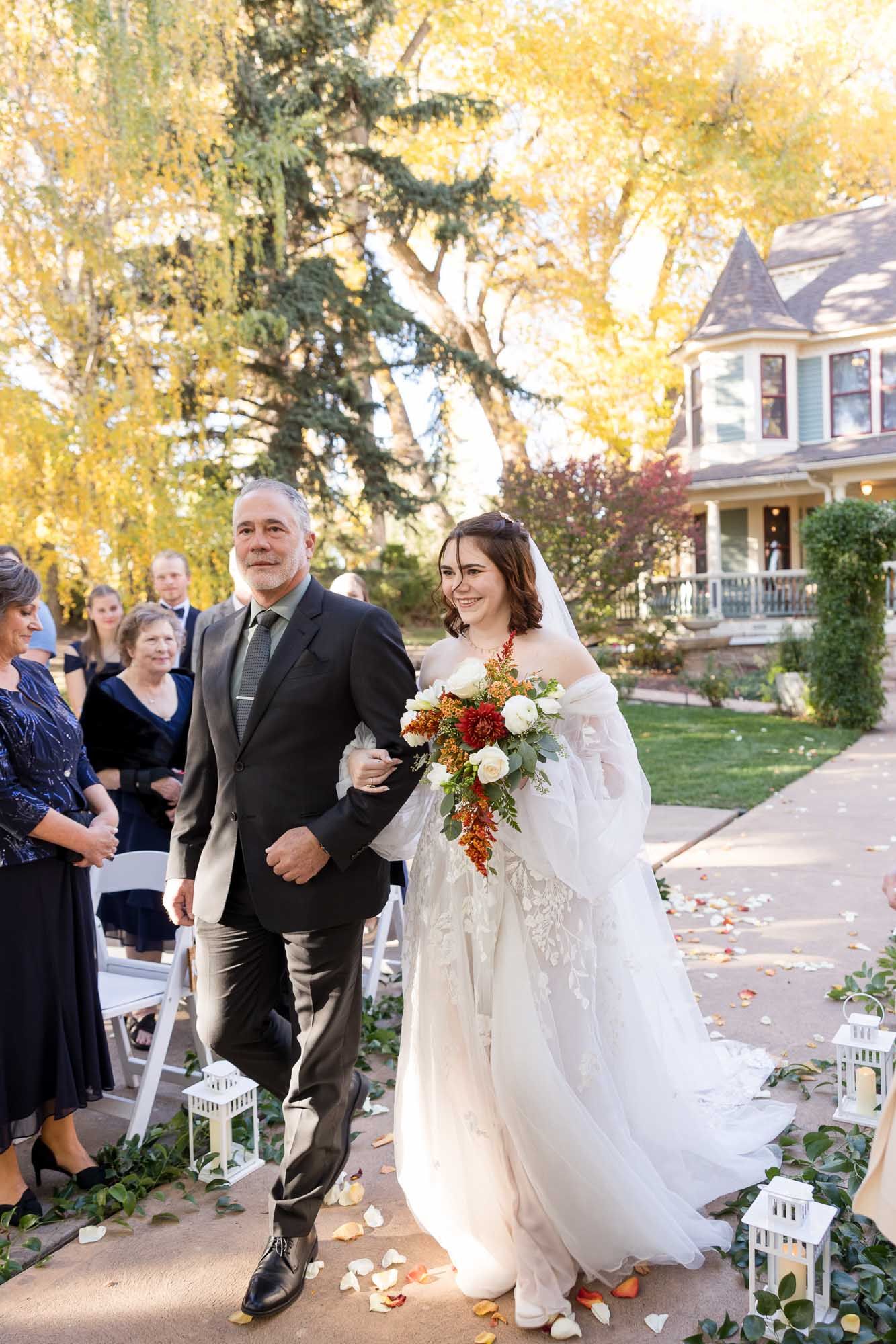 A wedding ceremony takes place under a draped pergola in an outdoor garden setting with autumn trees and scattered petals.