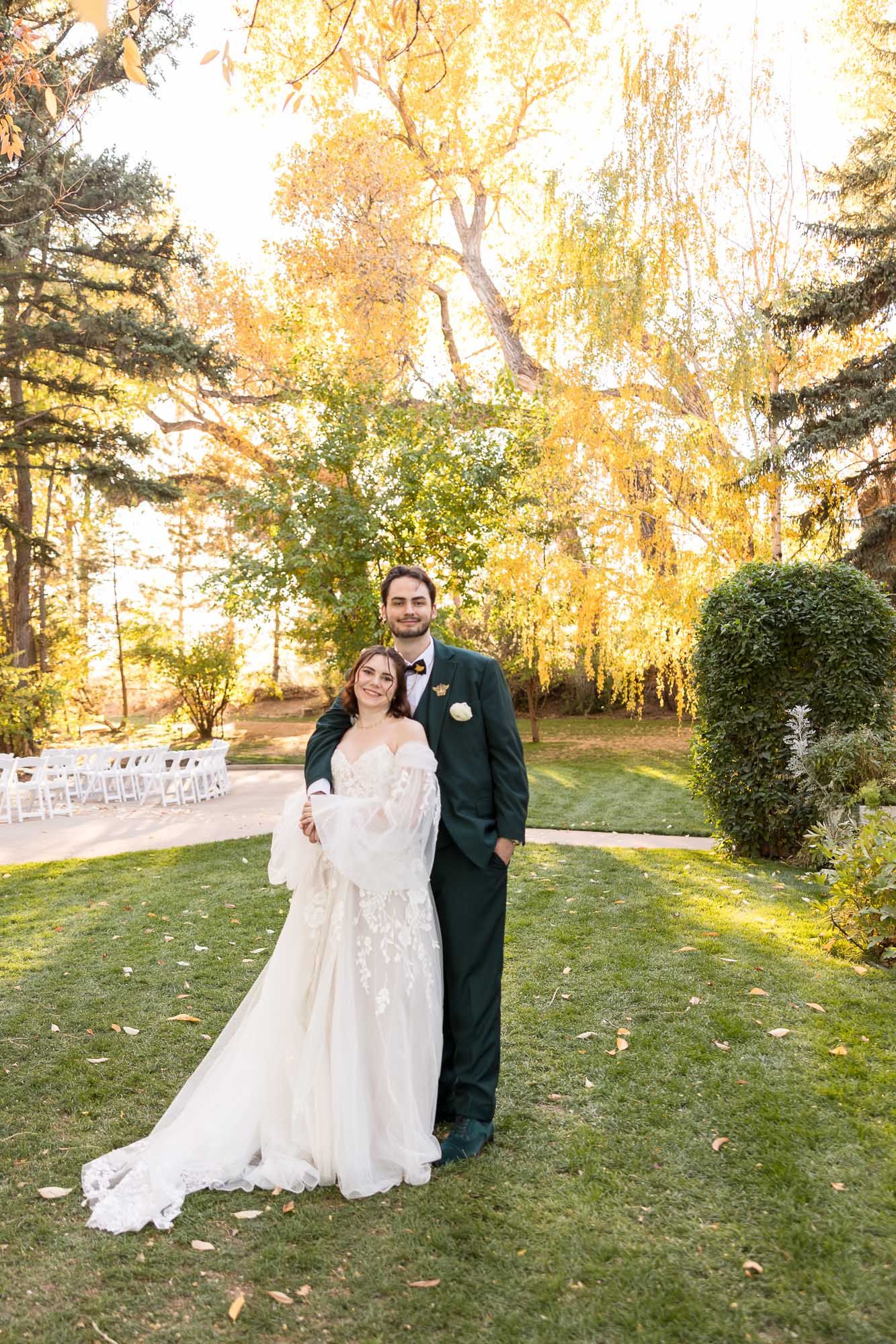A couple poses together outdoors on a grassy lawn with golden autumn trees glowing in the late afternoon sunlight.