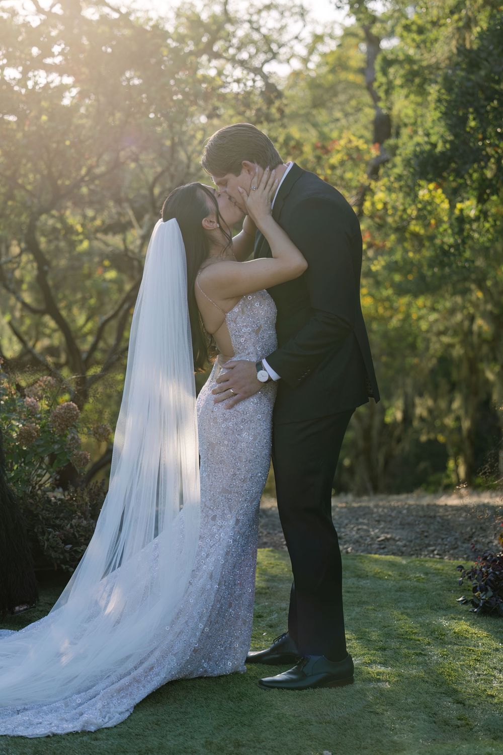 A couple shares an intimate moment in their wedding attire against a sunlit garden backdrop.
