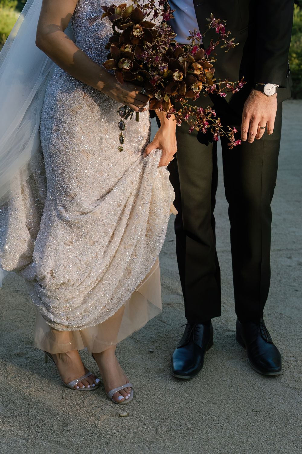 Close-up of bride in sparkly dress and groom's formal attire with burgundy floral bouquet.