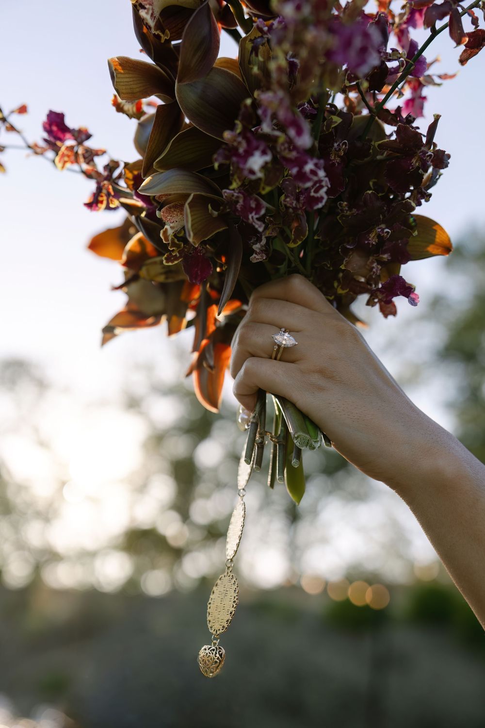 Hand holding a wedding bouquet with dark foliage and pink flowers against sunset sky.