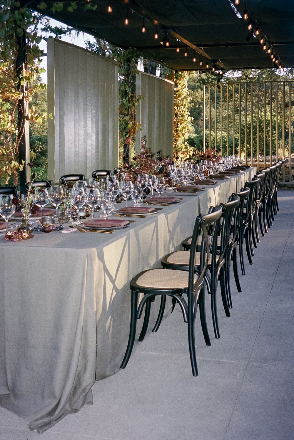Long dining tables set with elegant place settings and grey linens under string lights in an outdoor venue.