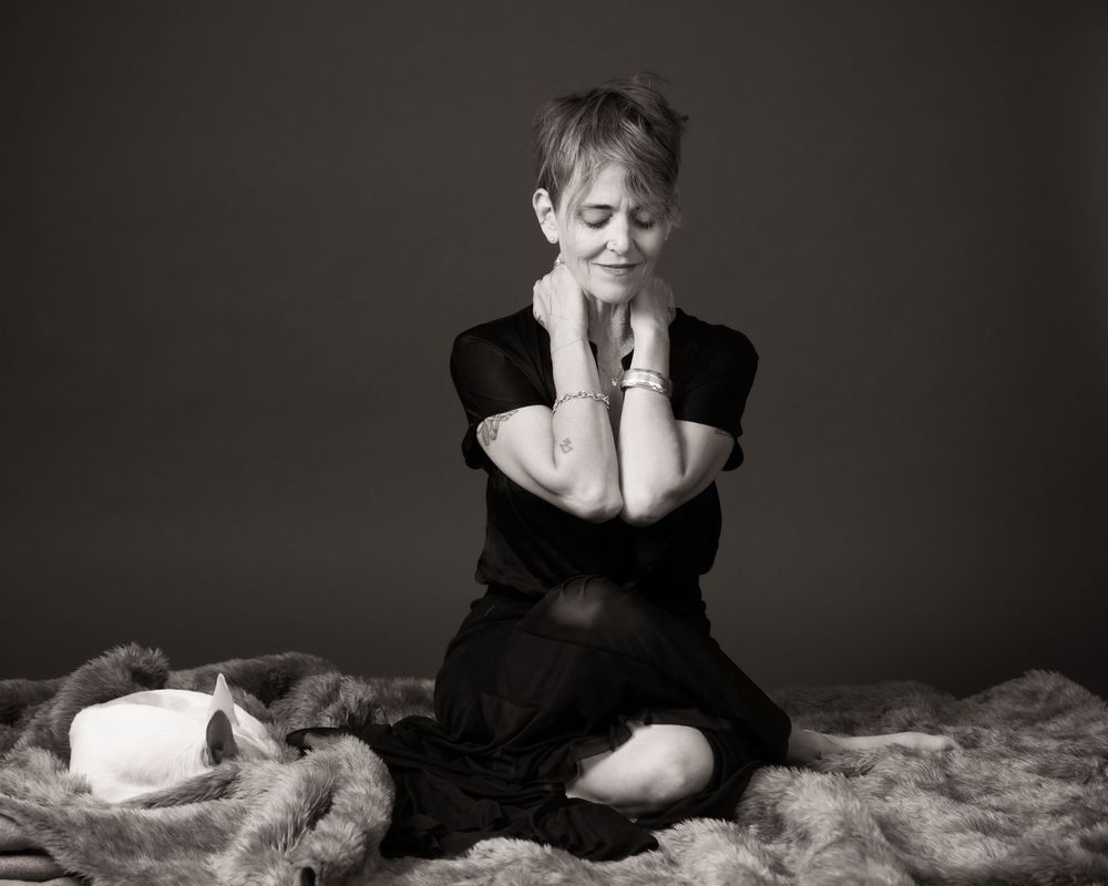 Black and white portrait of someone sitting on a textured blanket wearing dark clothing against a dark background.
