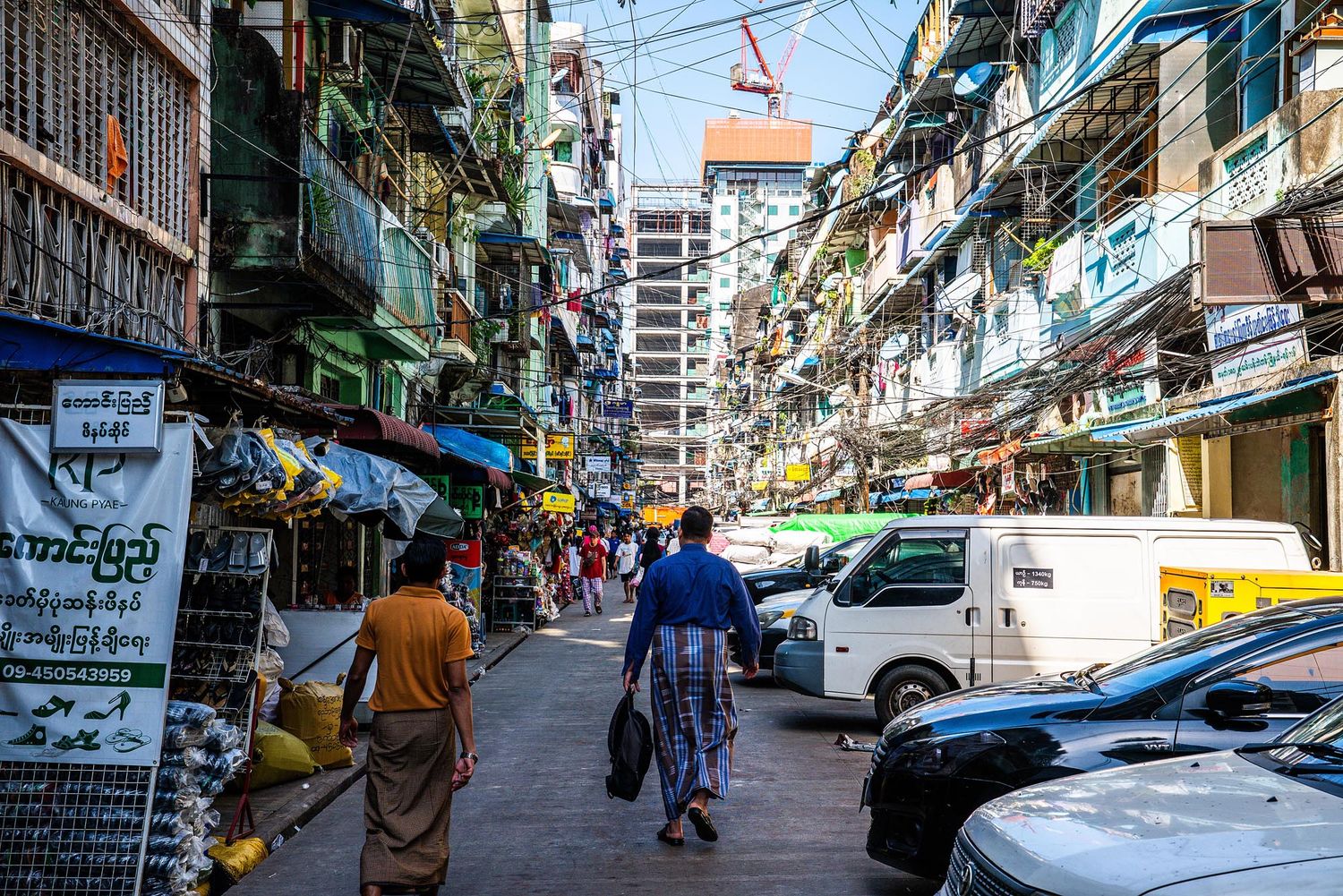 Narrow street scene in Yangon with tall residential buildings, parked vehicles, and people walking along the sidewalks.