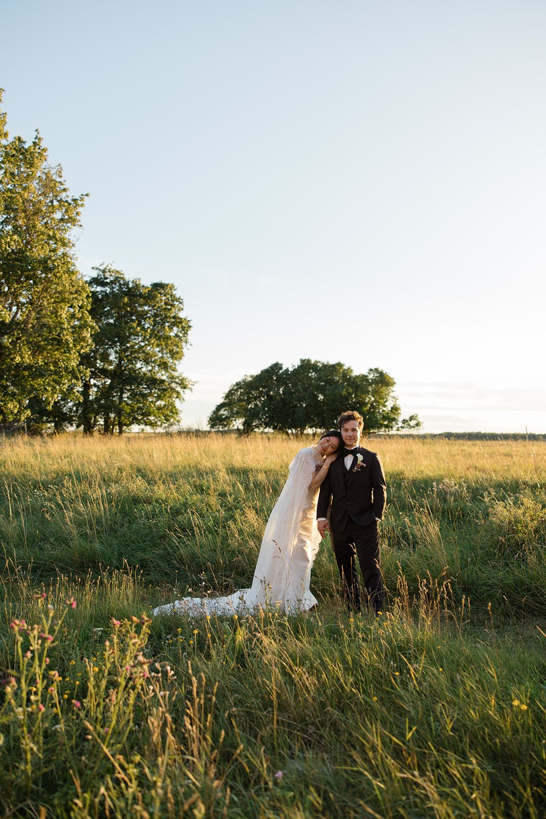 A romantic wedding couple shares a tender moment in a sunlit meadow with tall grass and trees at golden hour.