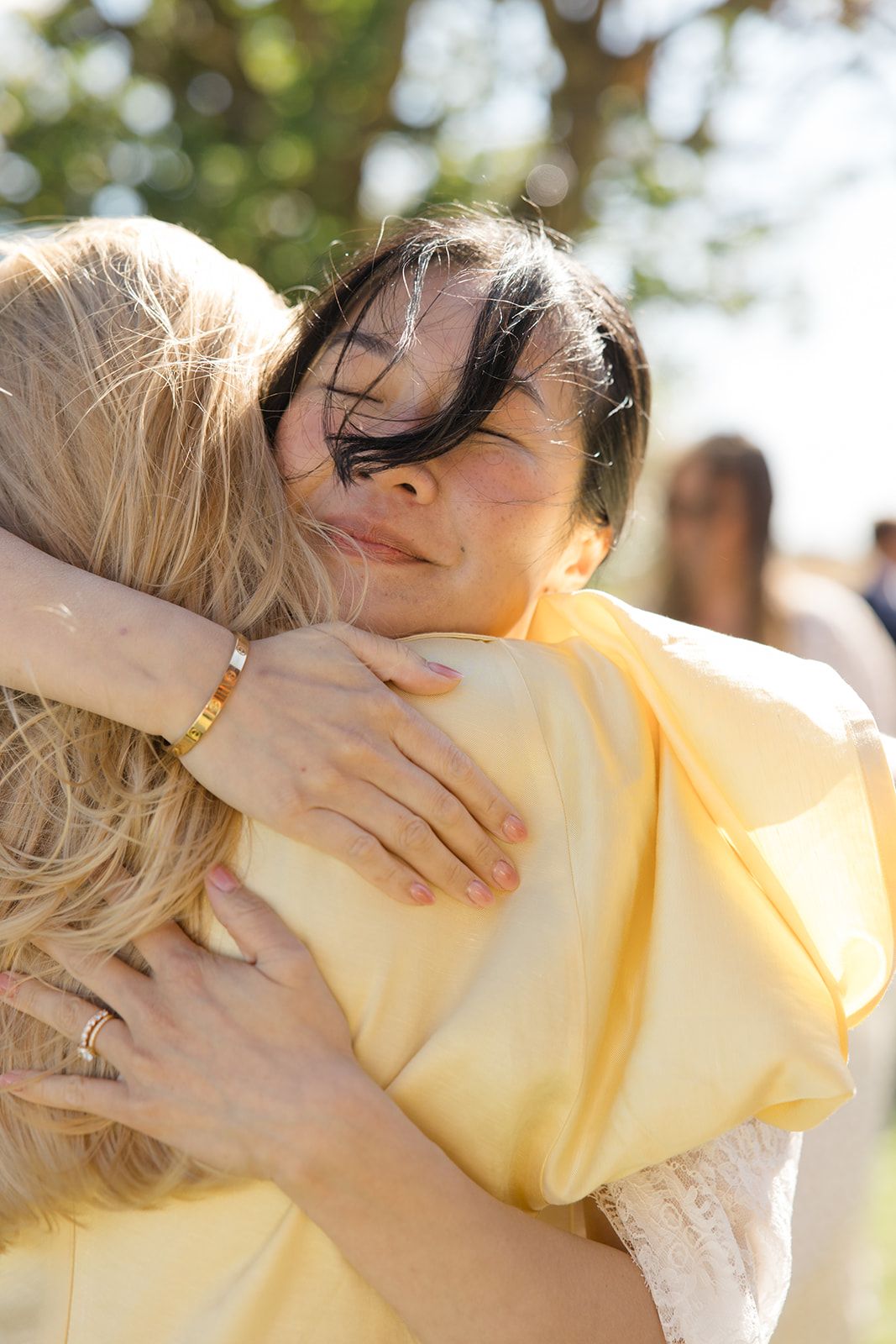 Two people share a warm embrace outdoors on a sunny day wearing light colored clothing.