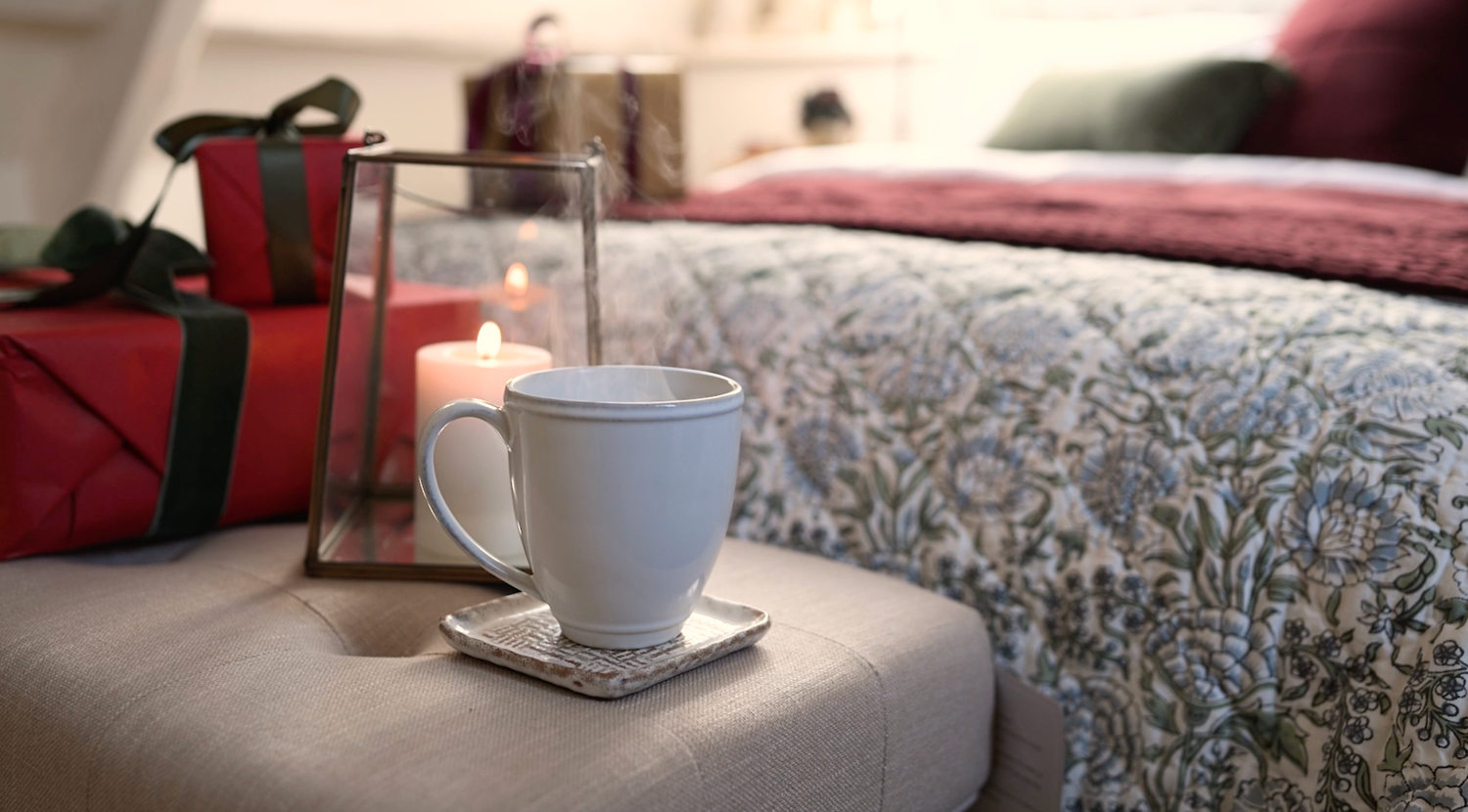 A mug of steaming hot tea sits at the end of a beautifully made bed with flickering candles and wrapped gifts