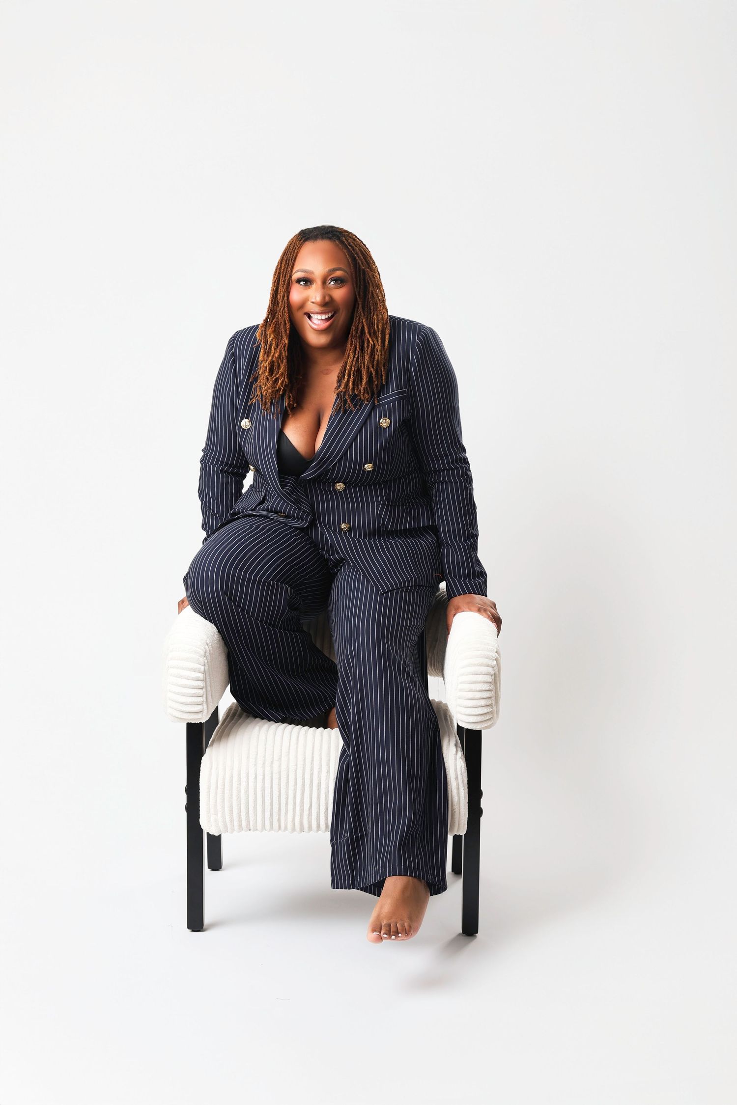 Professional wearing a navy pinstriped suit poses confidently in a white chair against a bright studio background.