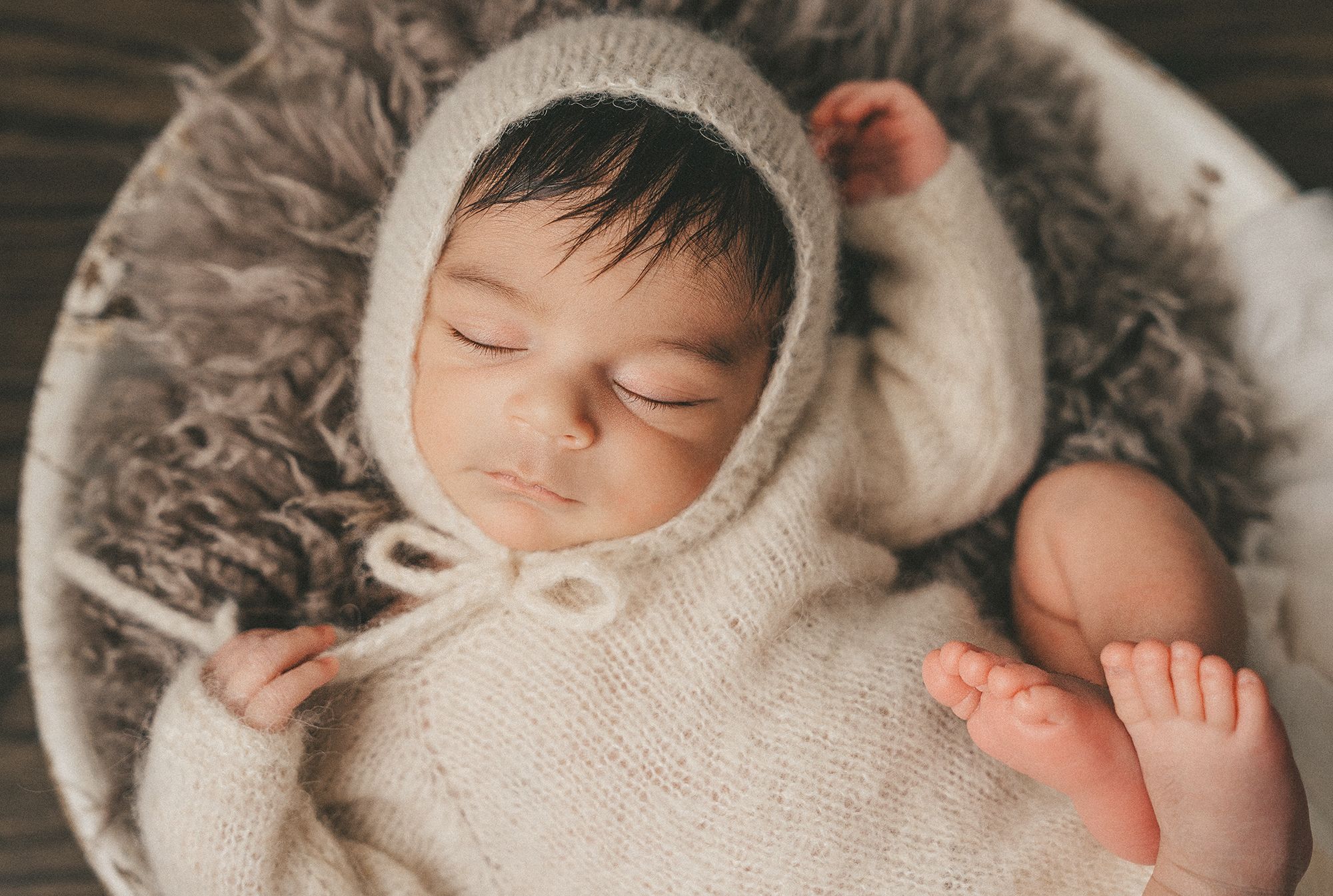 Sleeping newborn baby wearing white knitted bonnet rests peacefully on soft gray blanket.