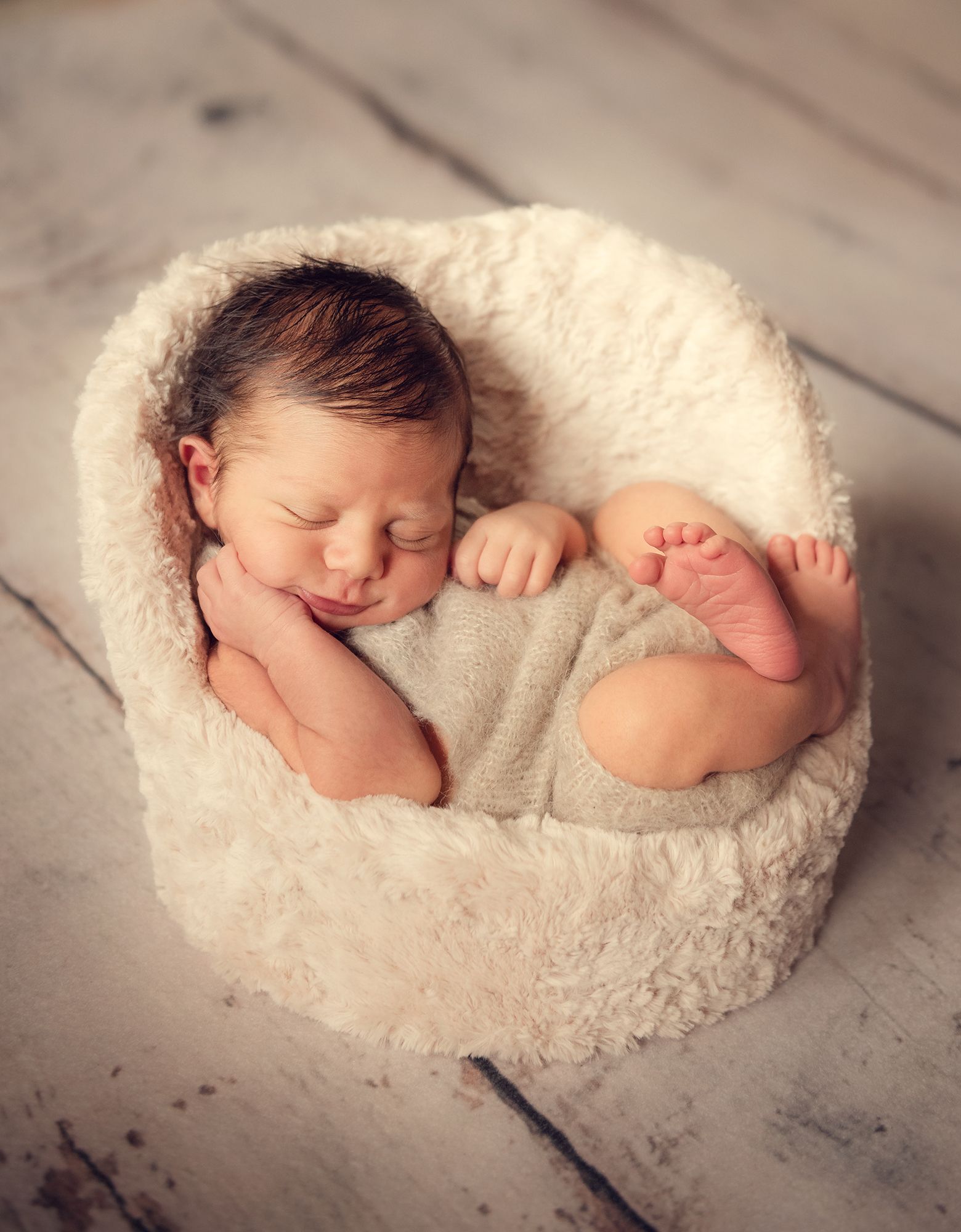 A sleeping newborn baby curled up on soft white fur blanket in a peaceful pose.