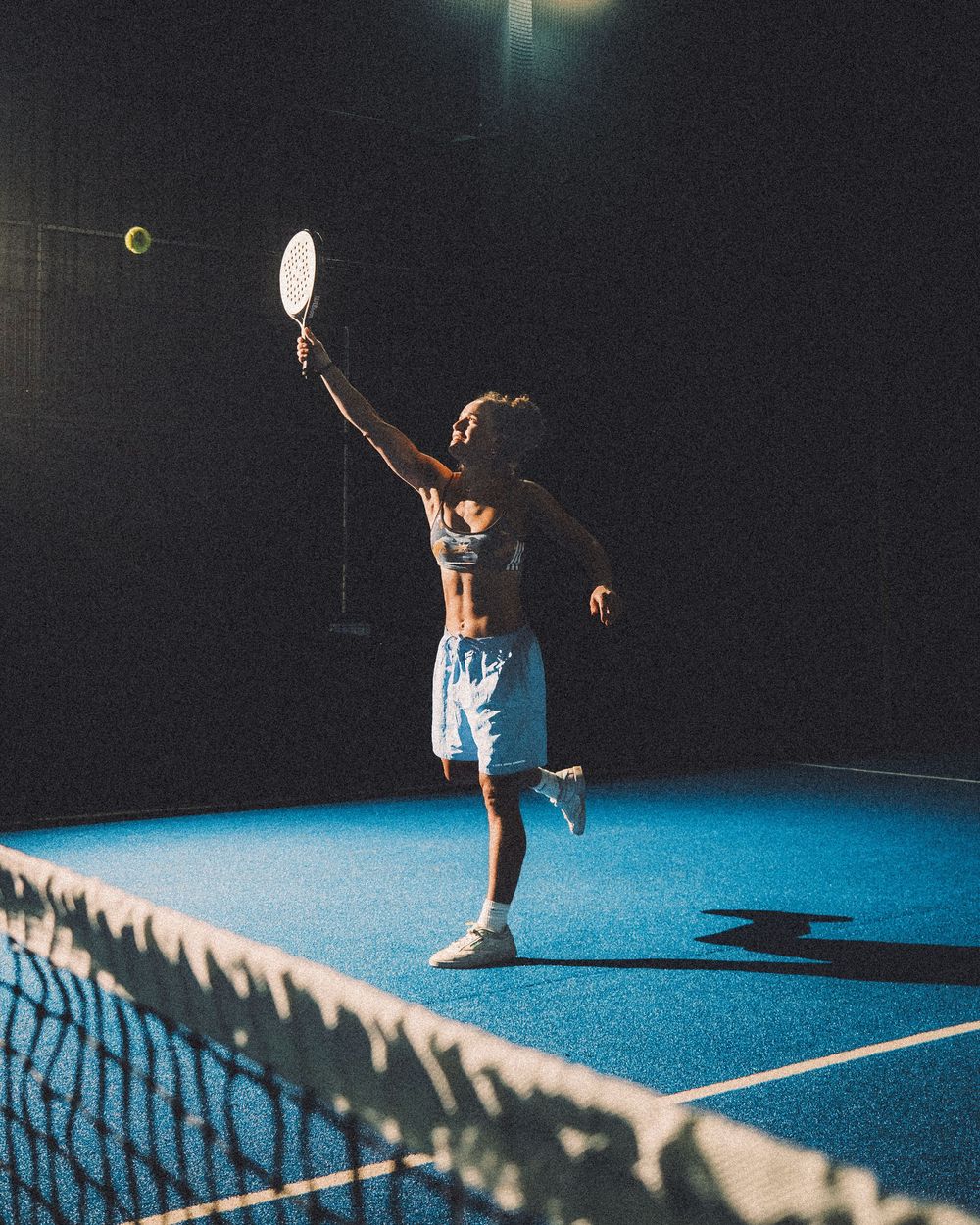 A tennis player leaps to serve under dramatic lighting on a blue court at night with a net in the foreground.