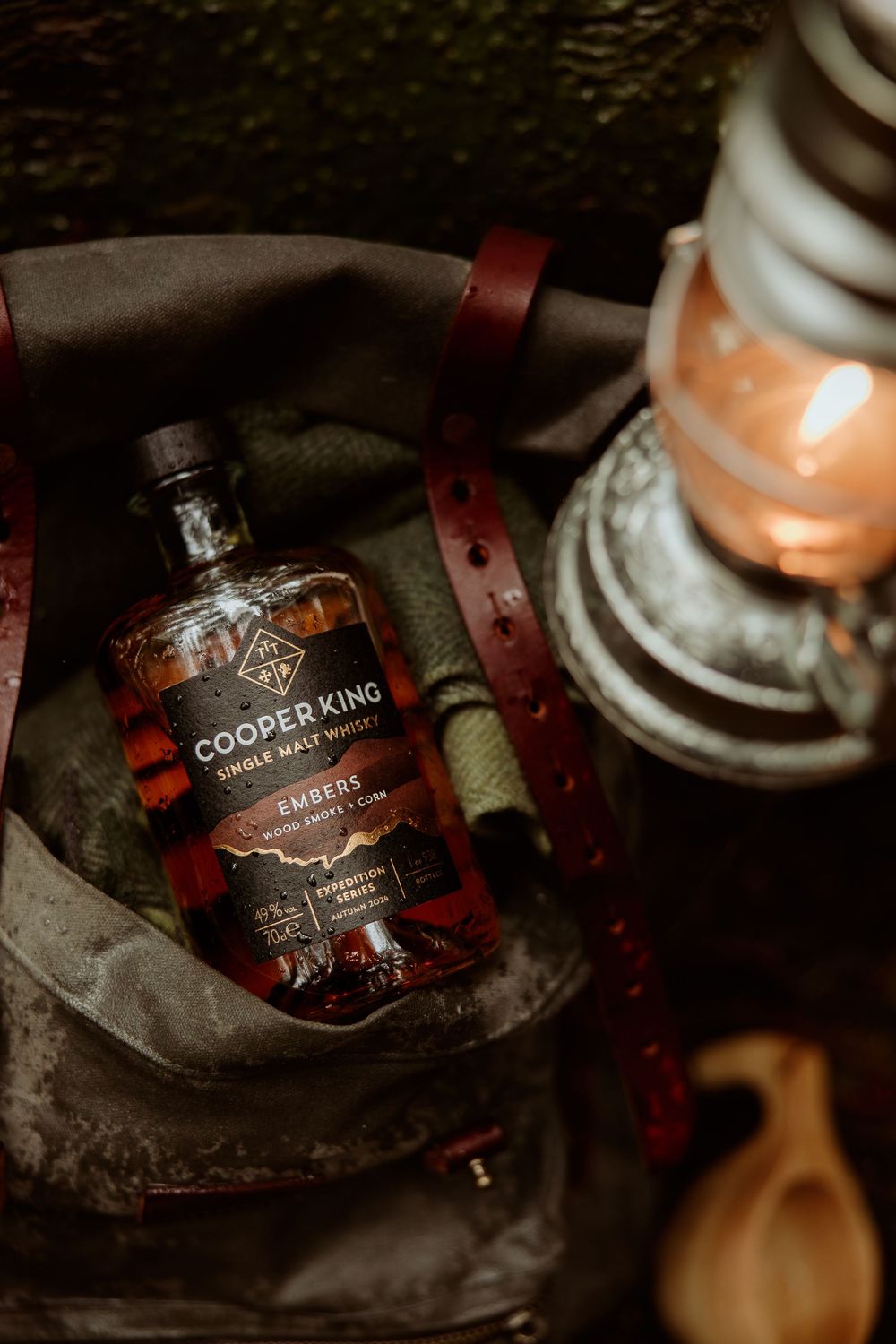 A bottle of Cooper King whiskey sits beside a vintage lantern and leather strap in a moody, dark setting.