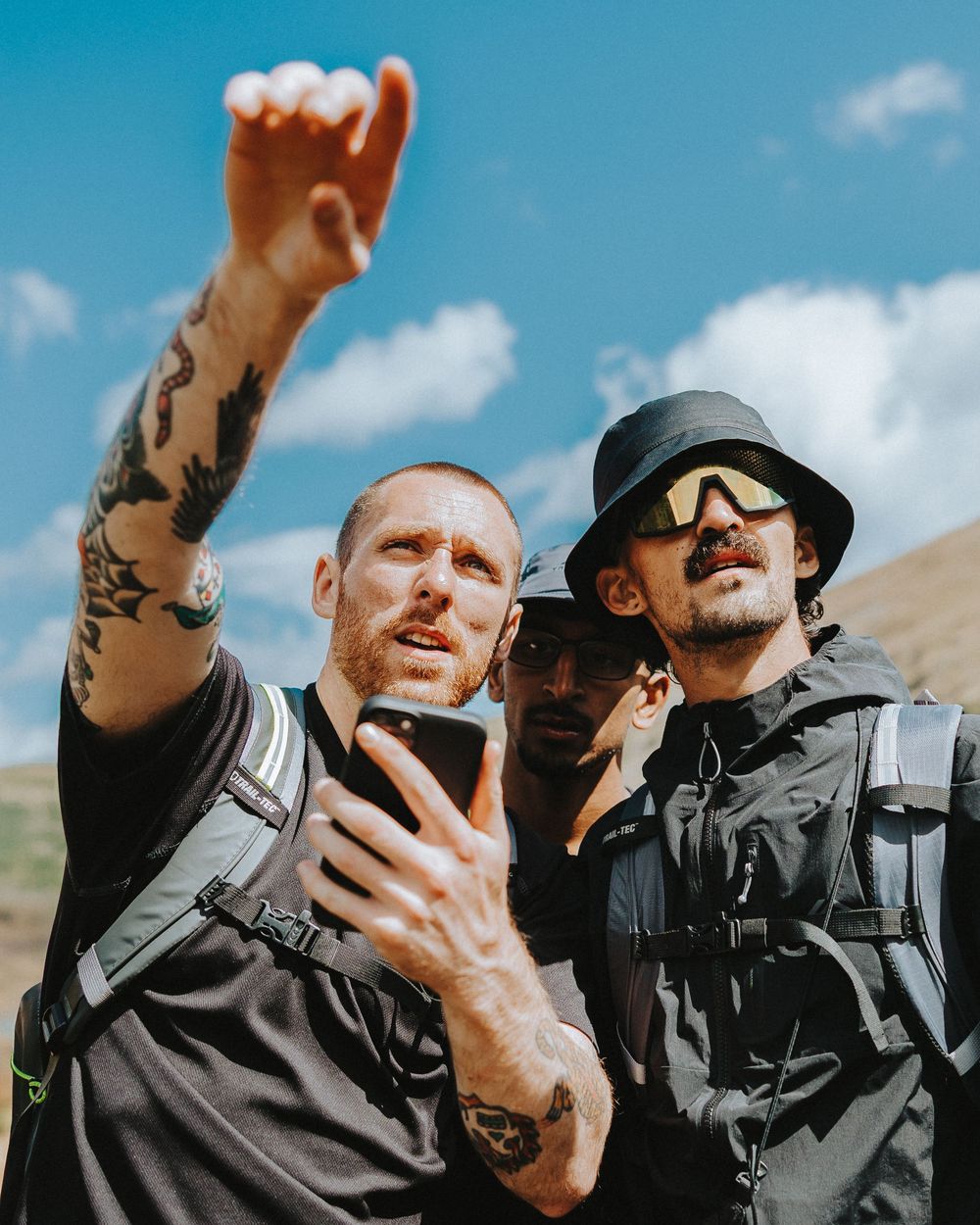 Two people in black tactical gear looking at a phone screen while pointing outdoors against a blue sky.