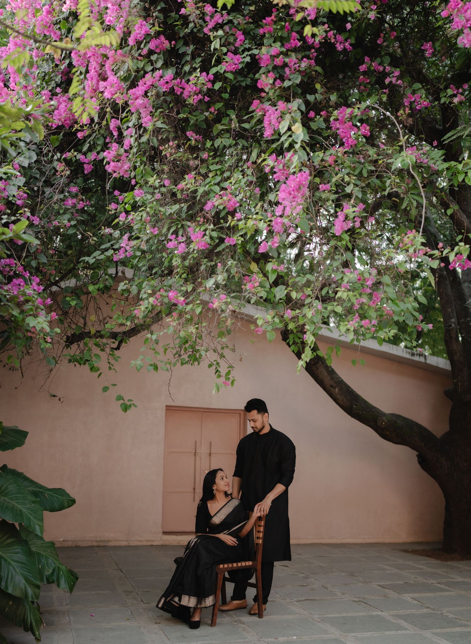 Couple in matching black outfits sharing a romantic moment under a blooming pink bougainvillea tree.