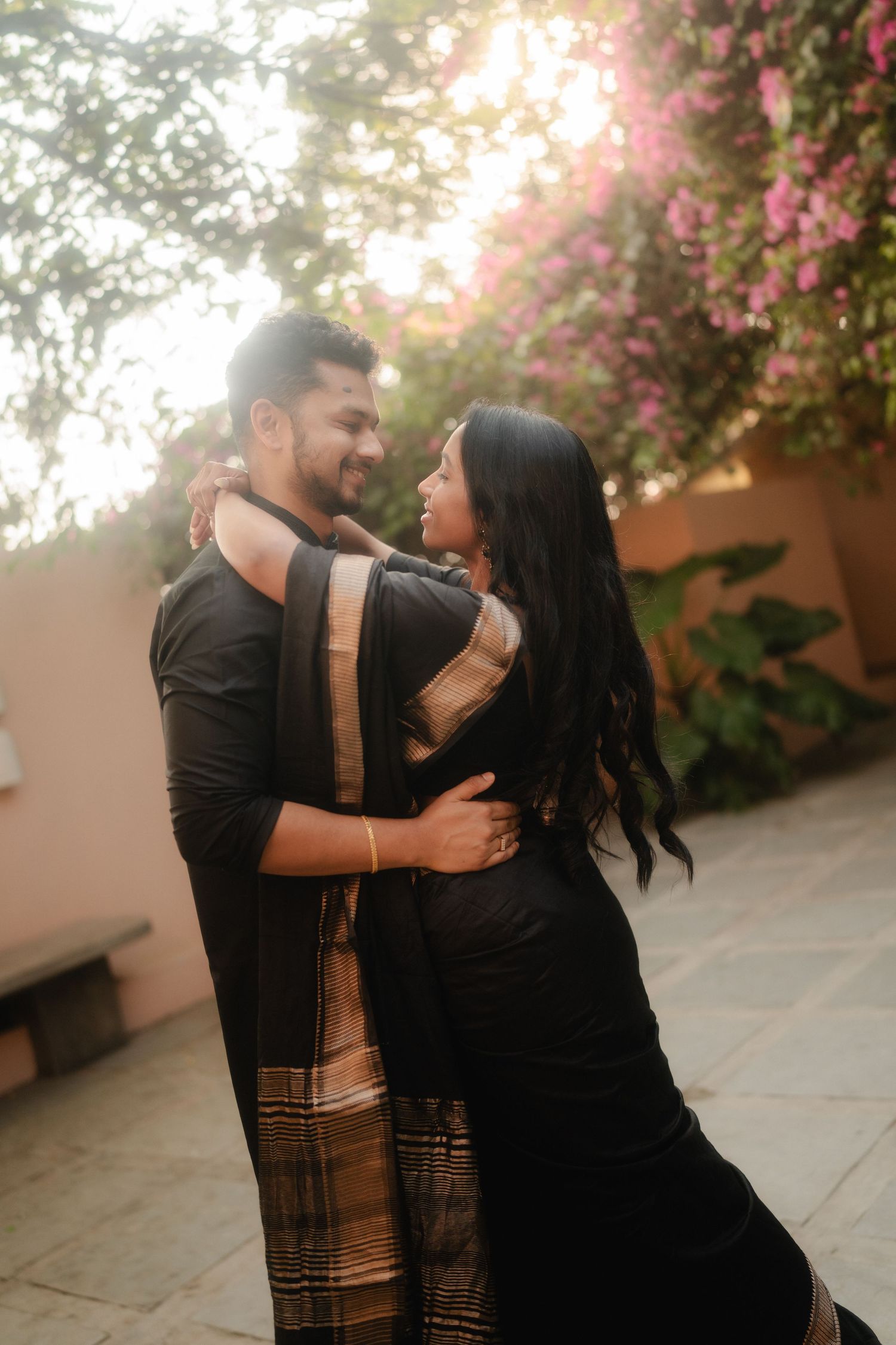 Romantic couple embraces outdoors against a backdrop of pink flowering trees at sunset.