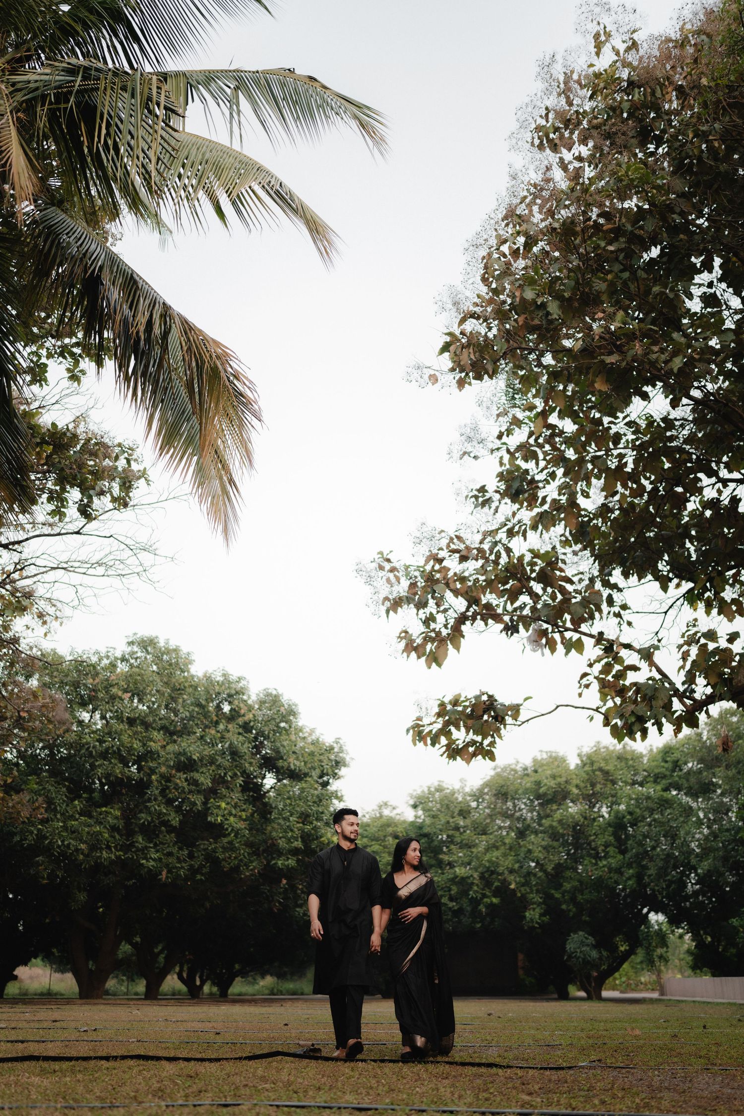 A couple dressed in matching black outfits stand together in a lush green park surrounded by tall trees.