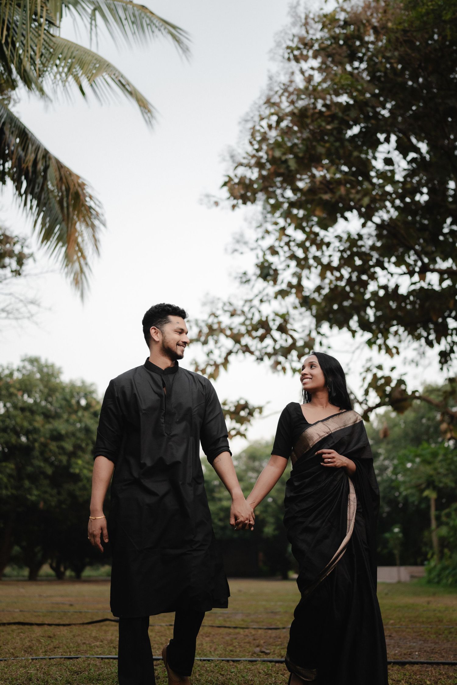 Couple in matching black traditional Indian attire holding hands in a lush green park setting.