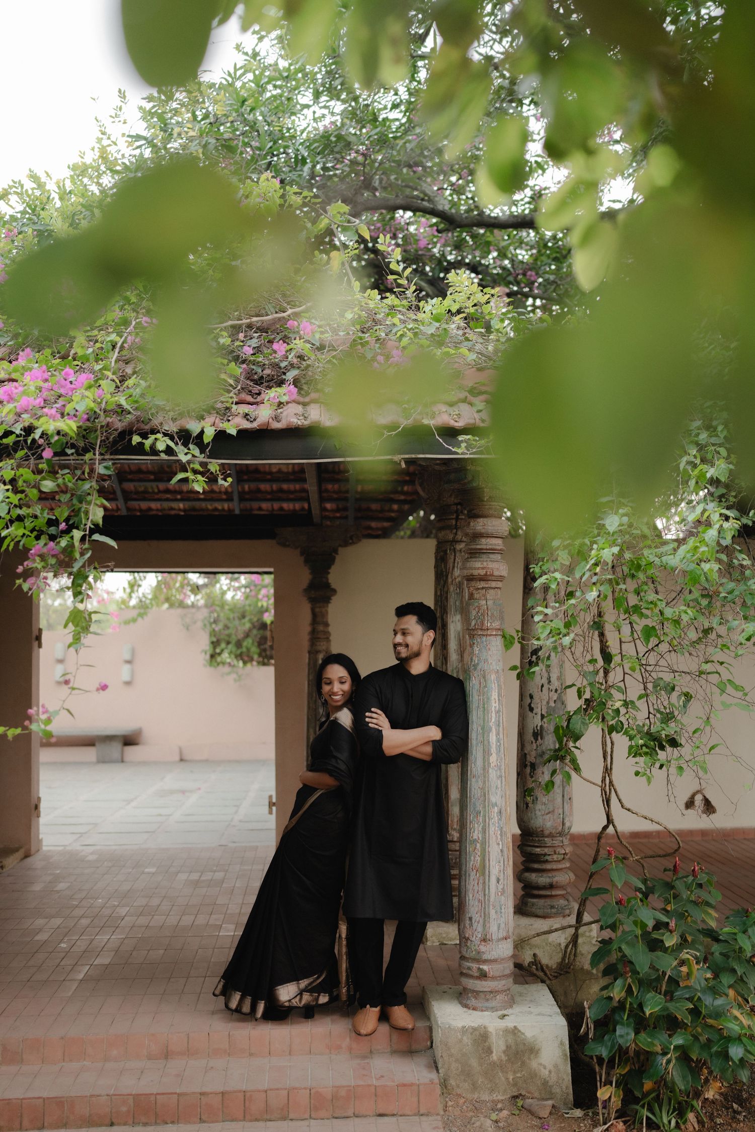 Couple in matching black traditional Indian attire posing under a stone pavilion surrounded by lush greenery and bougainvillea.